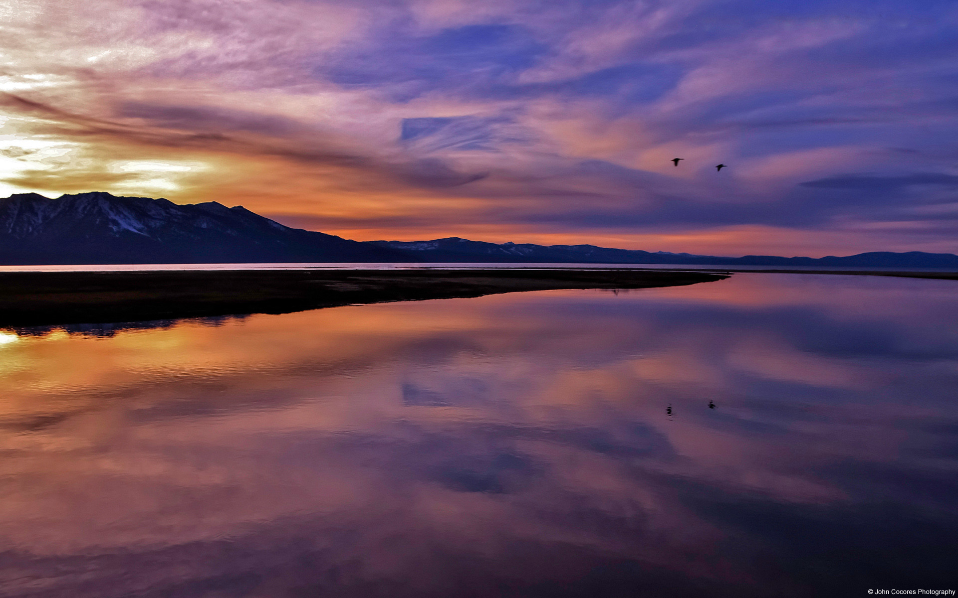 A breathtaking sunset over a serene lake with mountains in the background, reflecting the vibrant colors of the sky. Two birds fly in the distance, adding to the tranquil scene