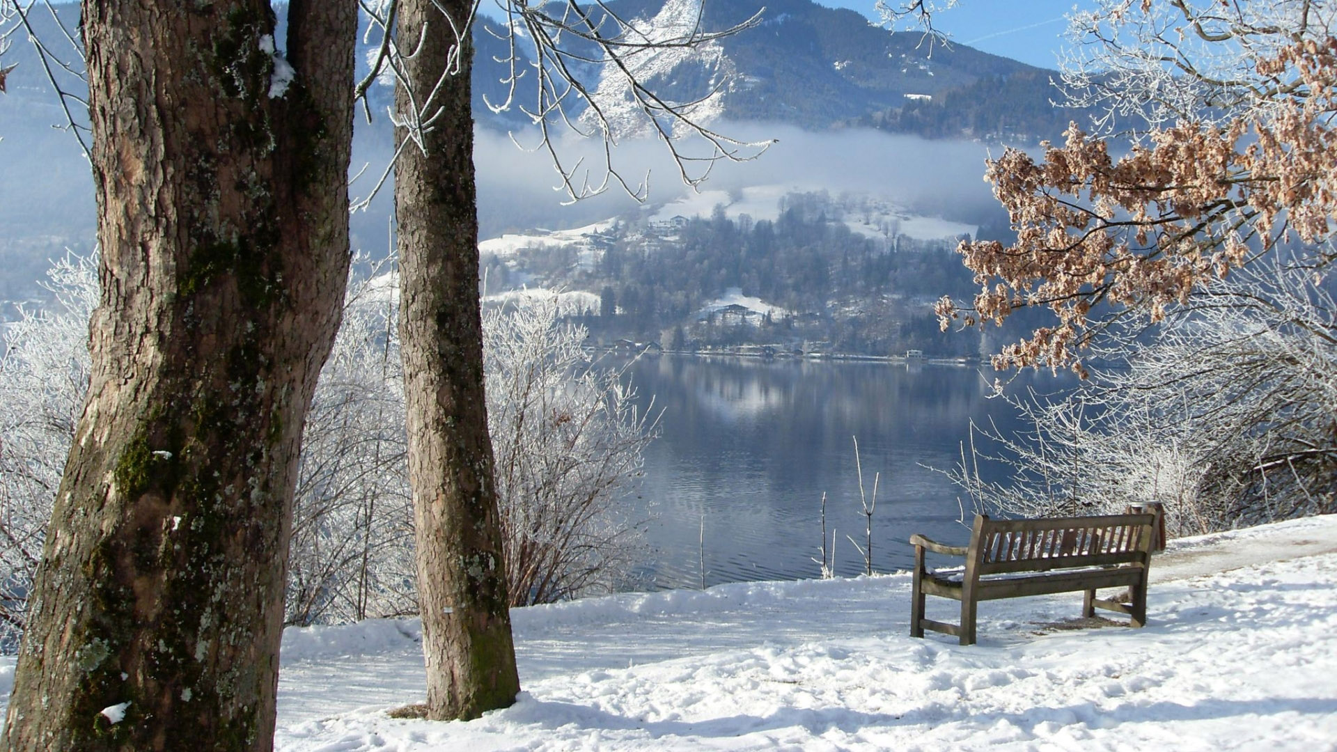 A serene winter landscape featuring a snow-covered bench overlooking a calm lake, with frost-covered trees and mountains in the background