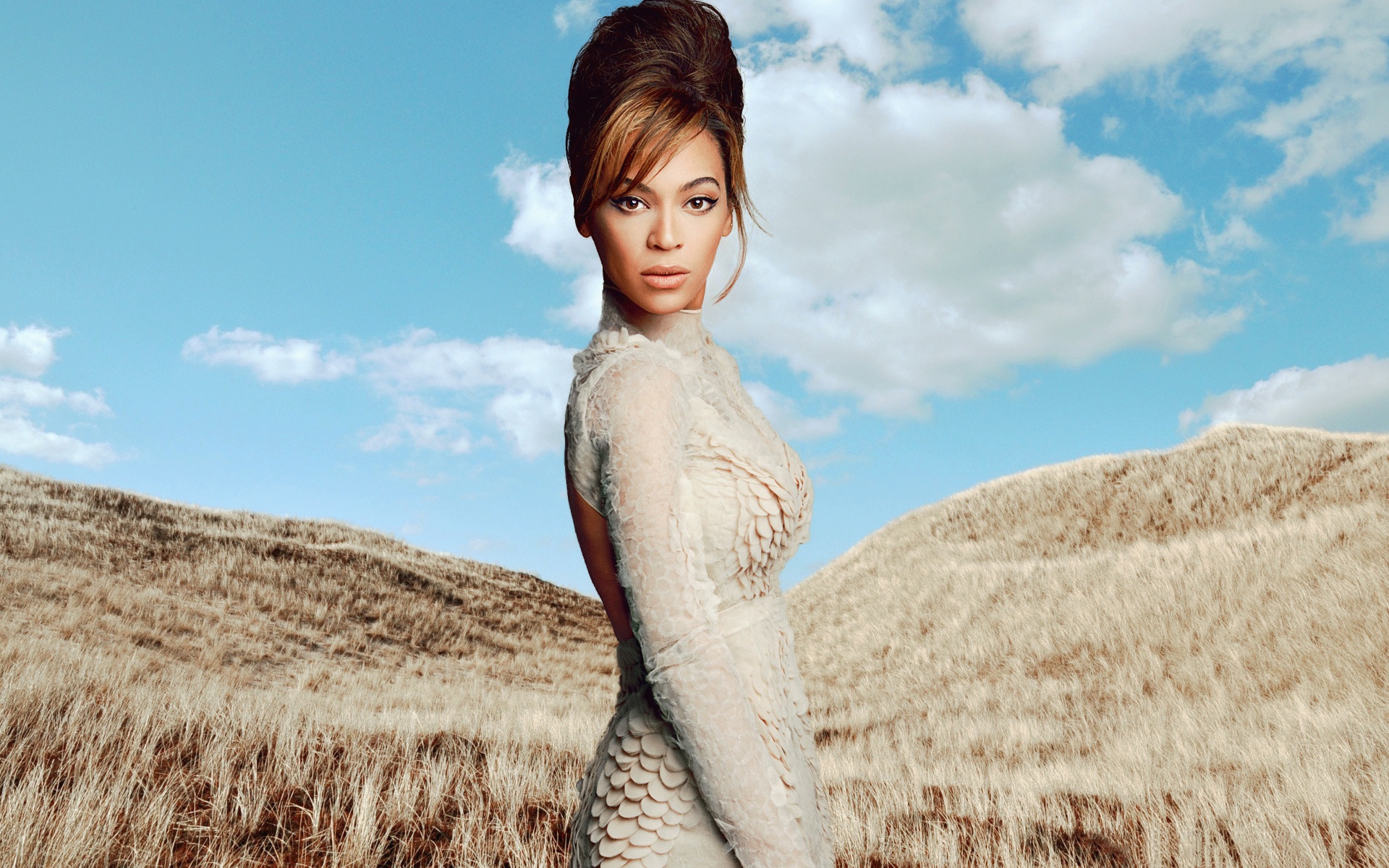 Beyoncé in a textured, light-colored gown, posing against a backdrop of rolling hills and a blue sky with scattered clouds