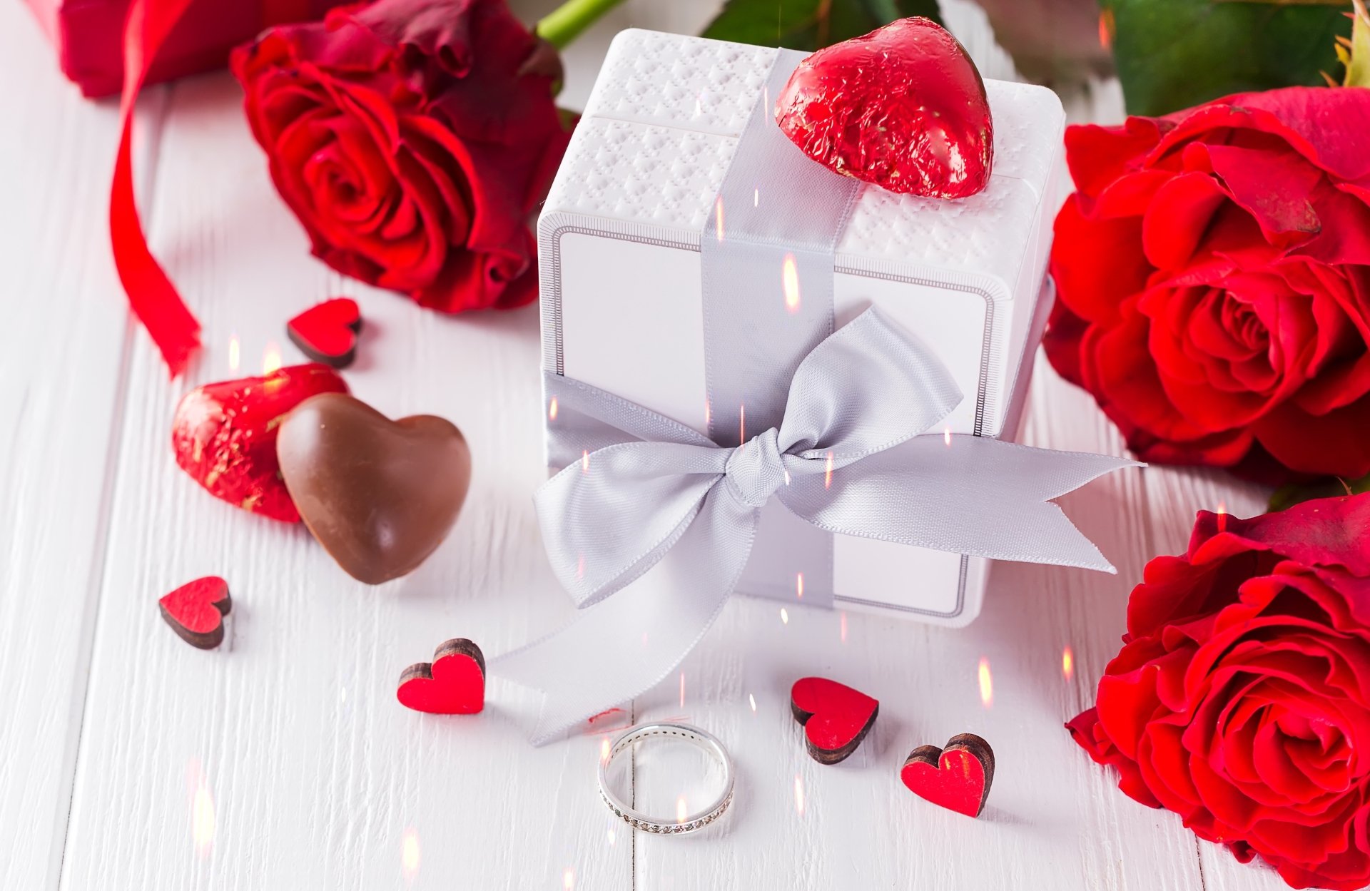 A Valentine's Day still life with red roses, a gift, chocolates, a ring, and heart-shaped decorations on a white wooden table