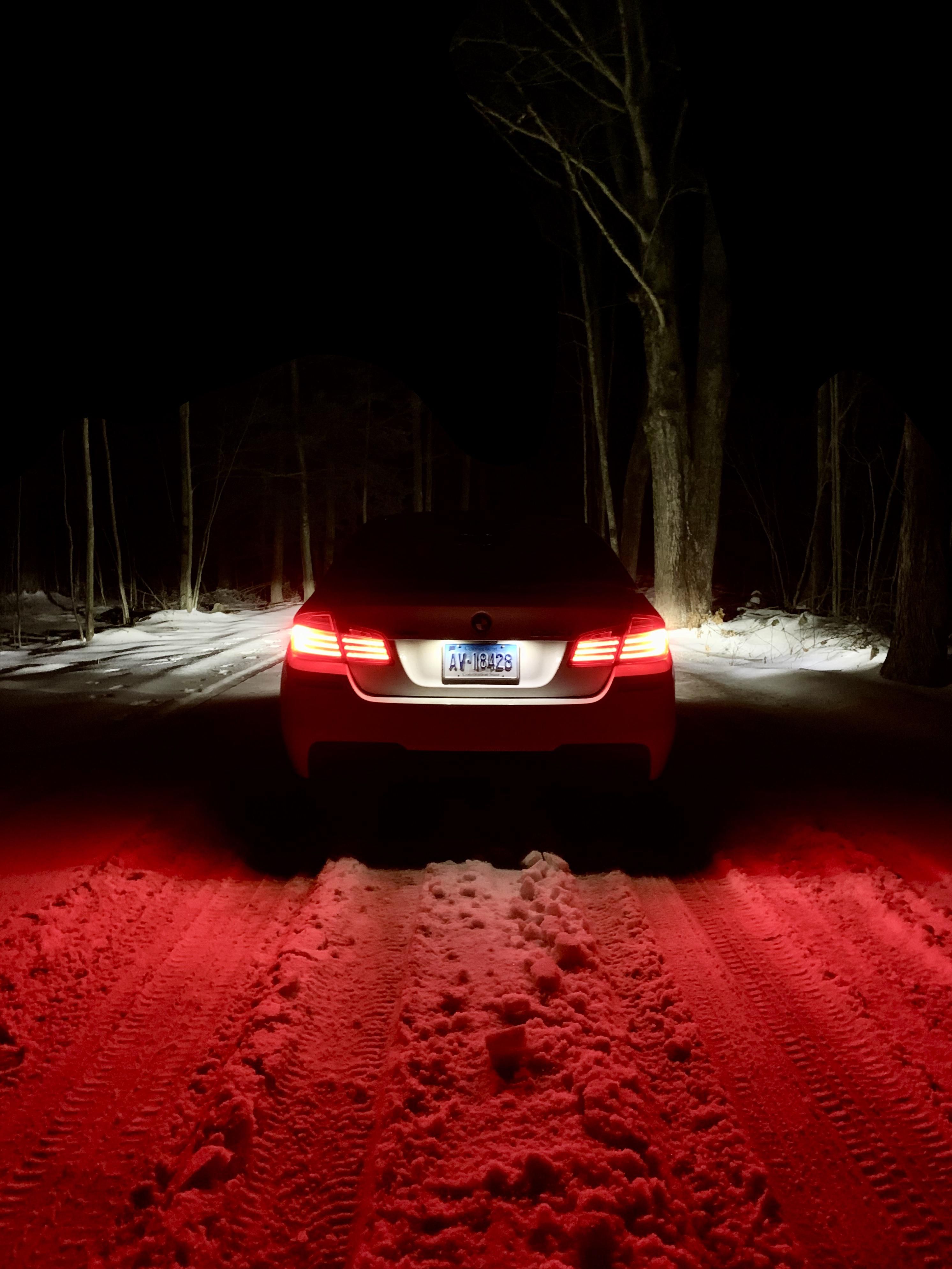 A BMW sedan with illuminated tail lights parked on a snow-covered road at night, surrounded by trees