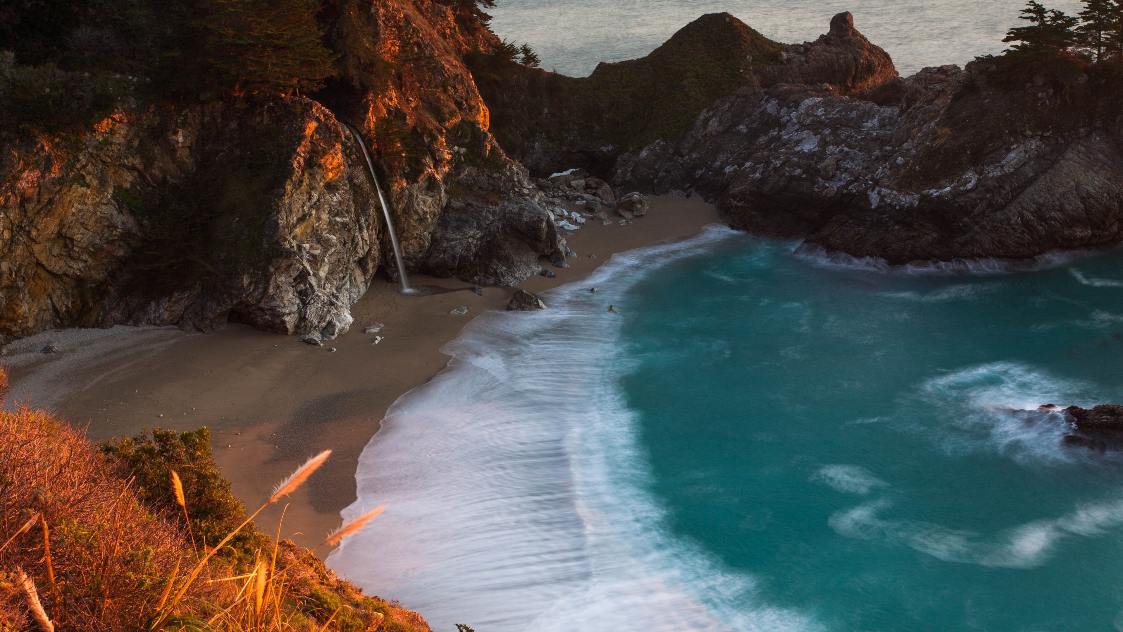 A breathtaking 4k wallpaper of McWay Falls in Julia Pfeiffer Burns State Park, California. A waterfall cascades onto a secluded beach with turquoise water and crashing waves, creating a stunning natural landscape