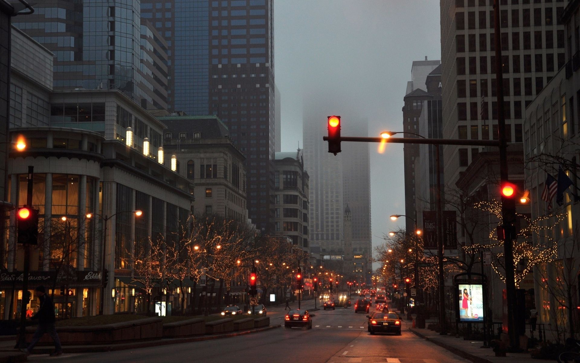 A cozy city street at dusk, illuminated by warm streetlights and the glow of traffic. The buildings are tall and imposing, but the twinkling lights and the soft fog create a sense of warmth and intimacy