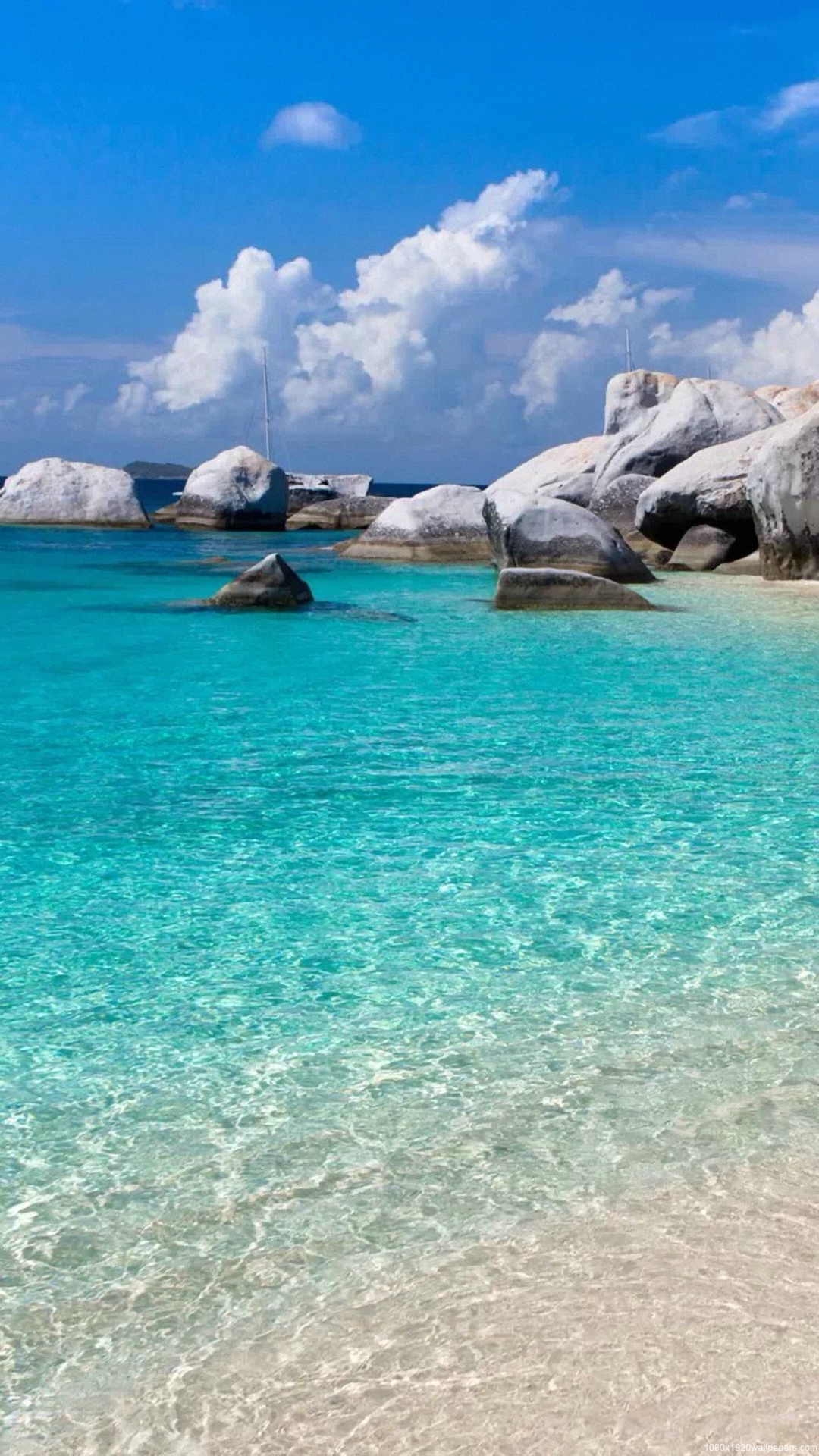 Tranquil turquoise waters gently lap against a pristine white sand beach, dotted with large granite boulders under a vibrant blue sky with fluffy white clouds. A sailboat rests in the calm bay, creating a serene tropical paradise scene