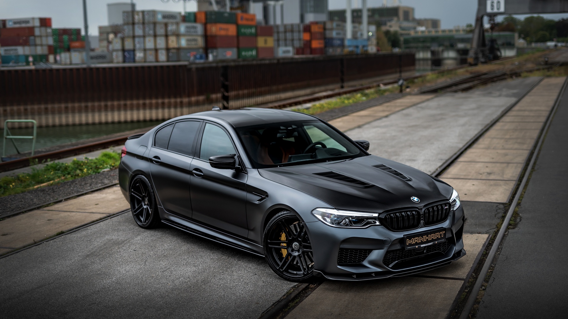 A matte gray BMW M5 sedan parked on a paved area near train tracks, featuring aftermarket modifications such as a black hood with vents and custom wheels