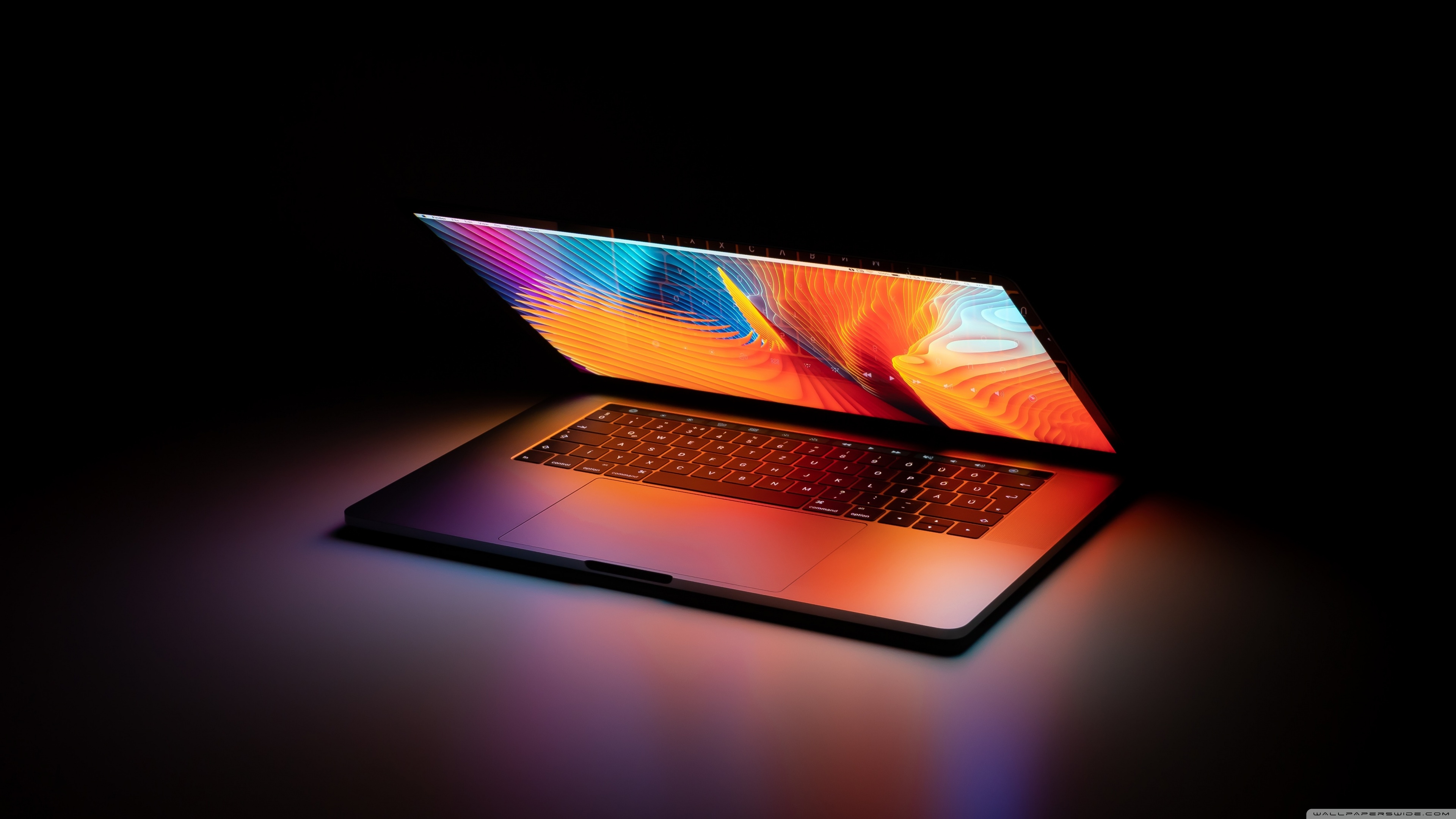 A sleek, modern laptop glows in a dark room, its screen displaying a vibrant abstract image. The warm light emanating from the display illuminates the keyboard and surrounding surface