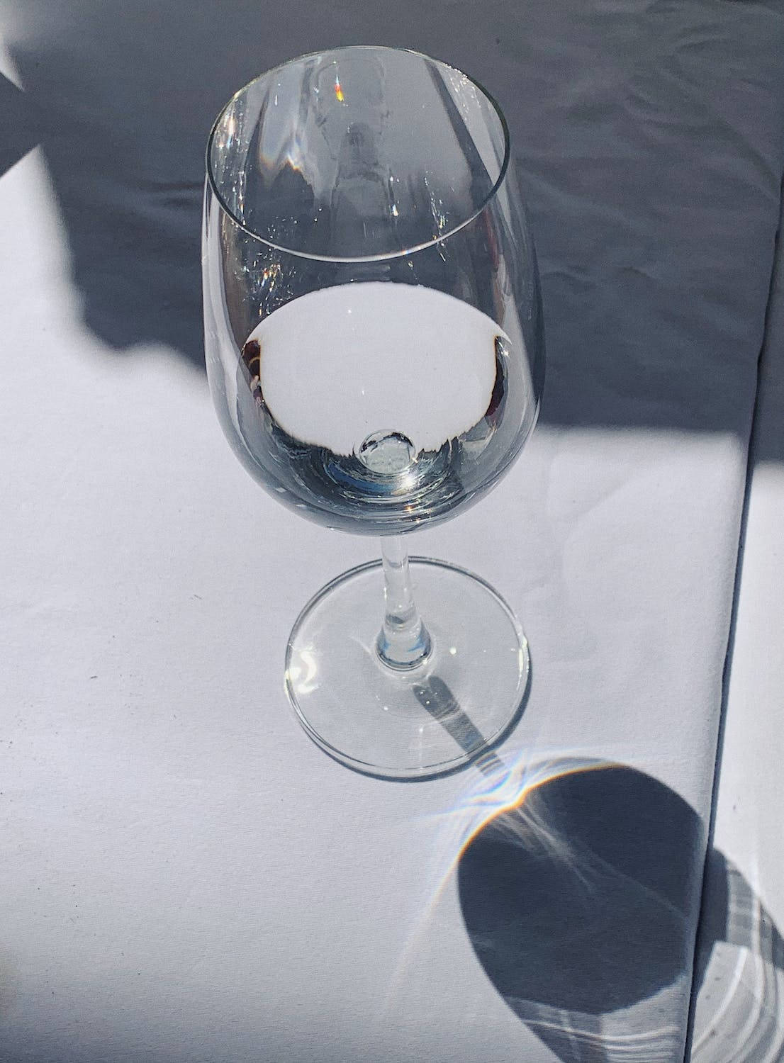 A clear wine glass sits on a white tablecloth, with sunlight creating a rainbow prism effect