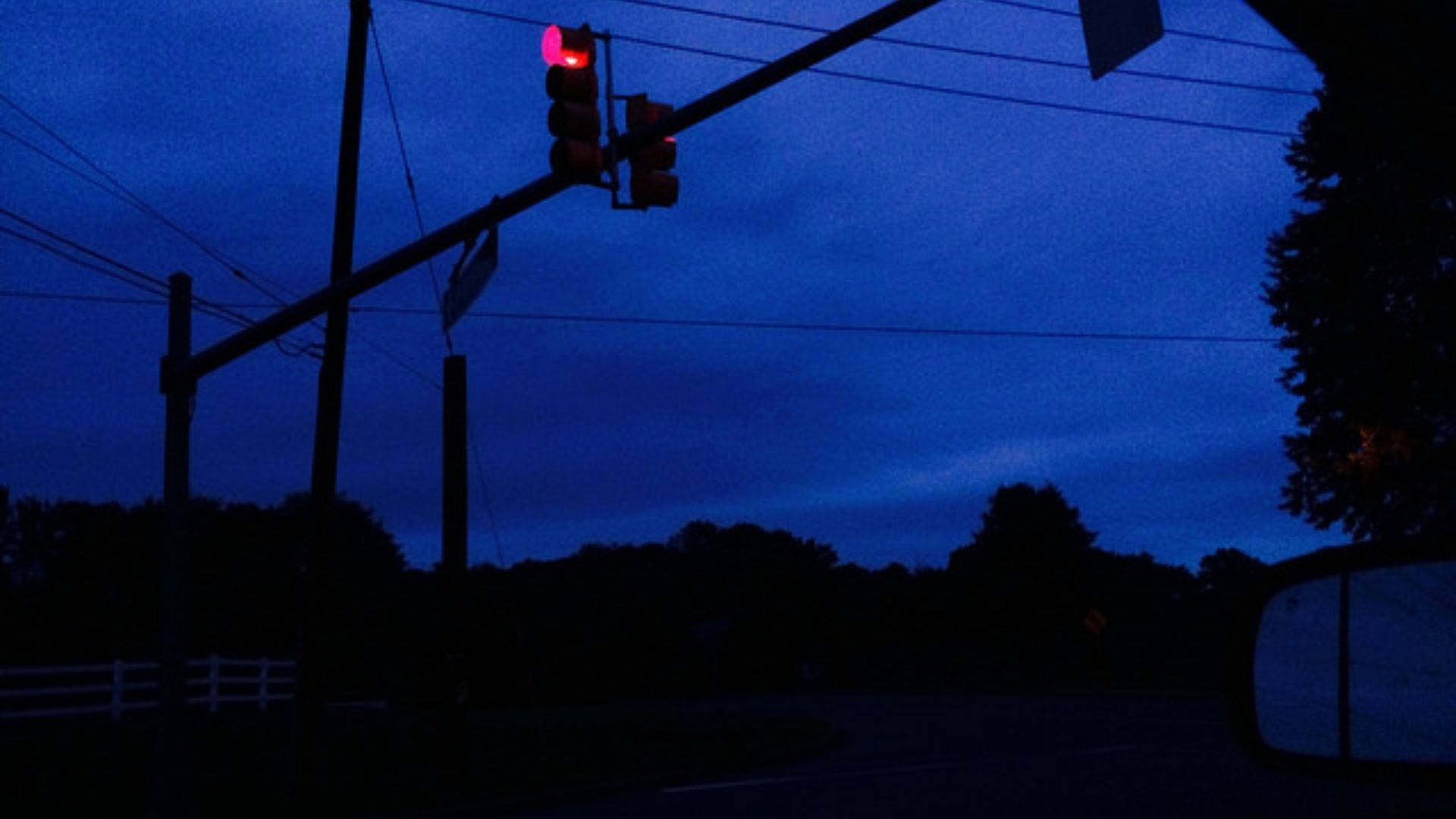 A neon-blue hued evening scene at a rural intersection, featuring a traffic light with a glowing red signal
