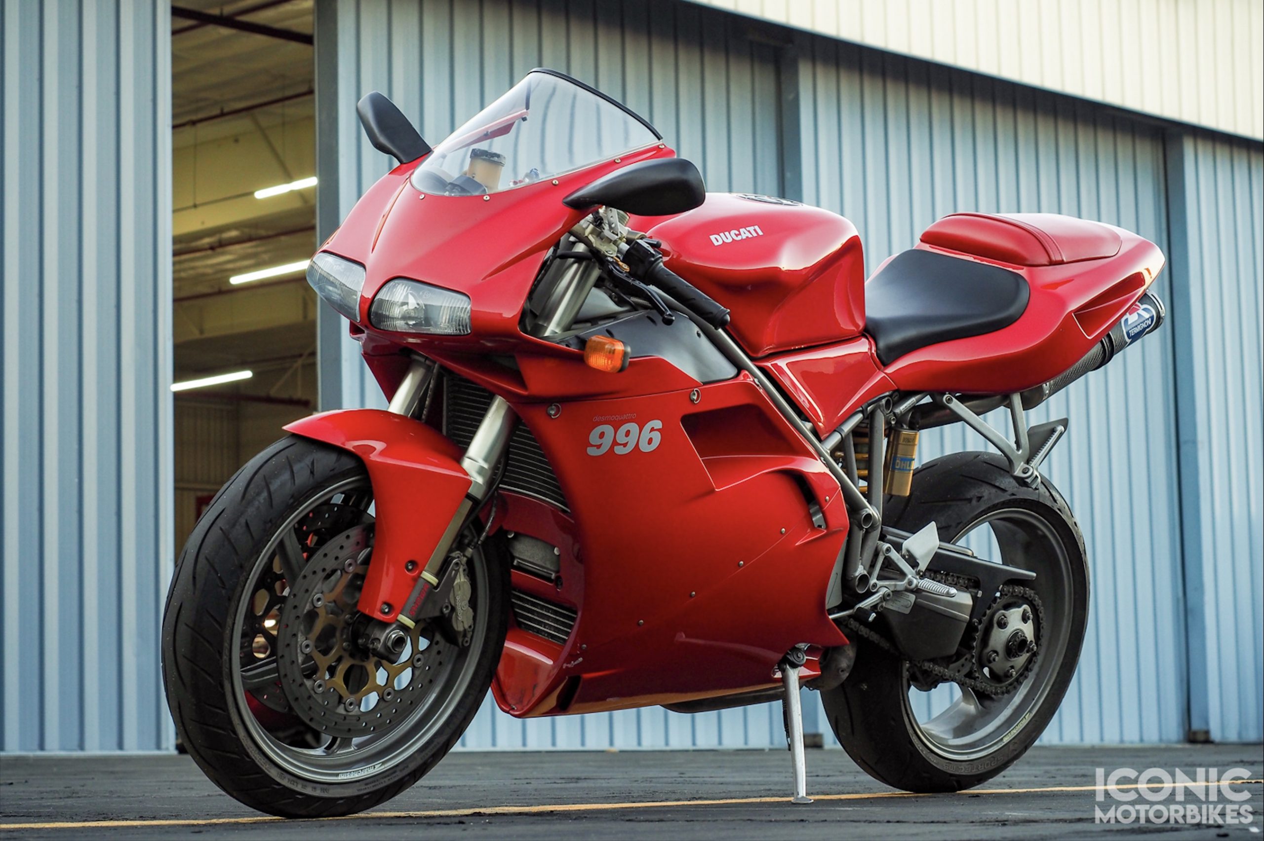 A Ducati 996 motorcycle parked with a backdrop of a corrugated metal wall. The bike is in profile, showcasing its vibrant red color, sleek design and powerful stance