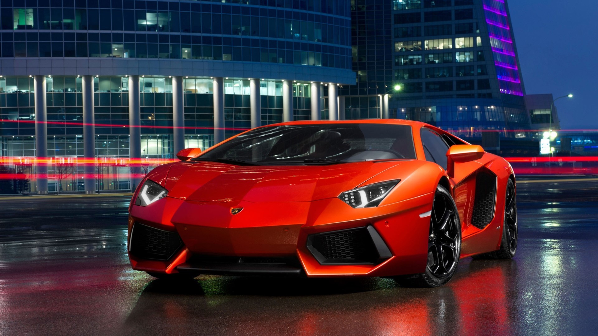 A vibrant orange Lamborghini Aventador parked in front of a modern building at night, reflecting in the wet pavement