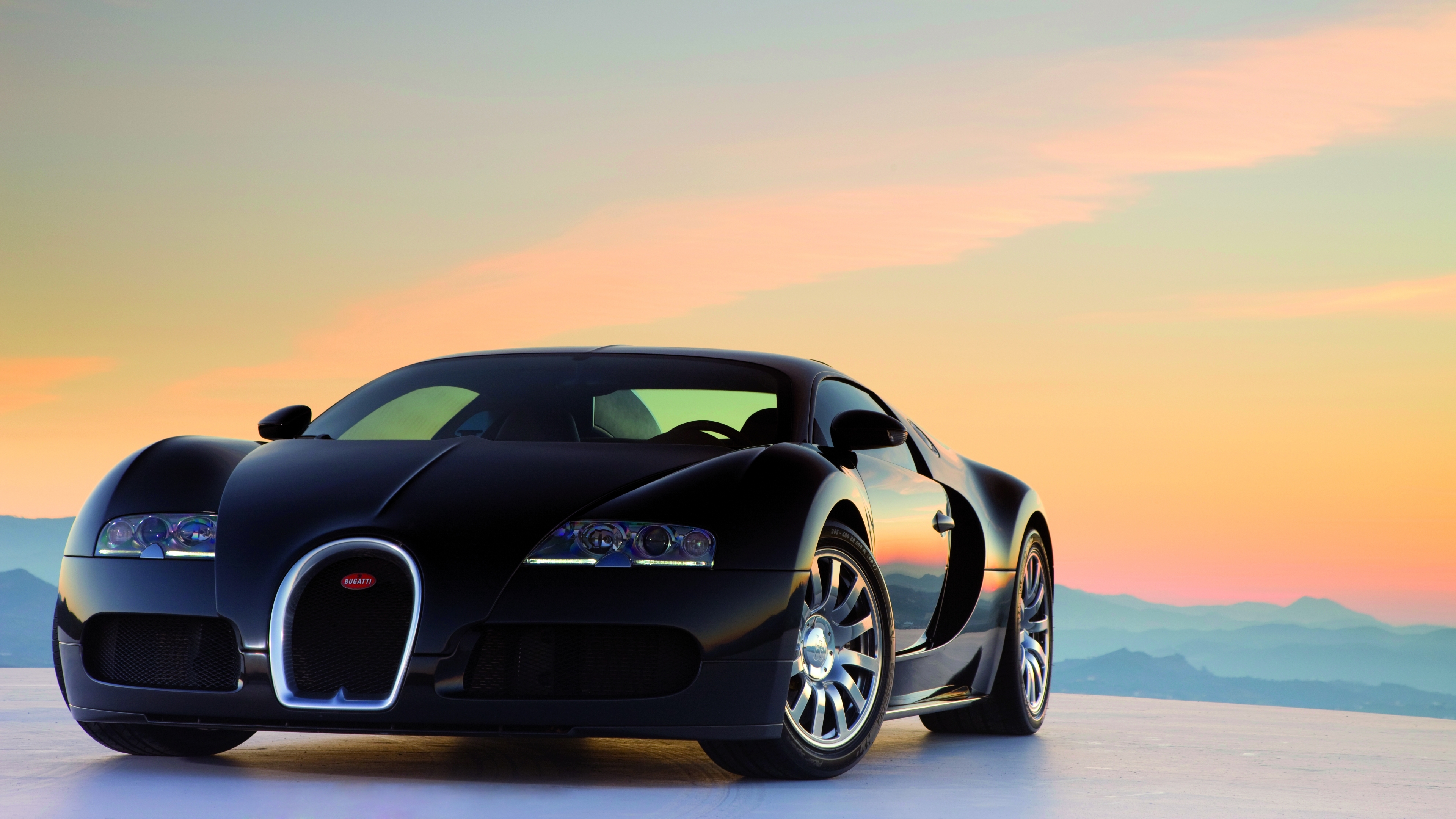 A sleek black Bugatti Veyron sports car parked against a backdrop of a beautiful sunset and mountains