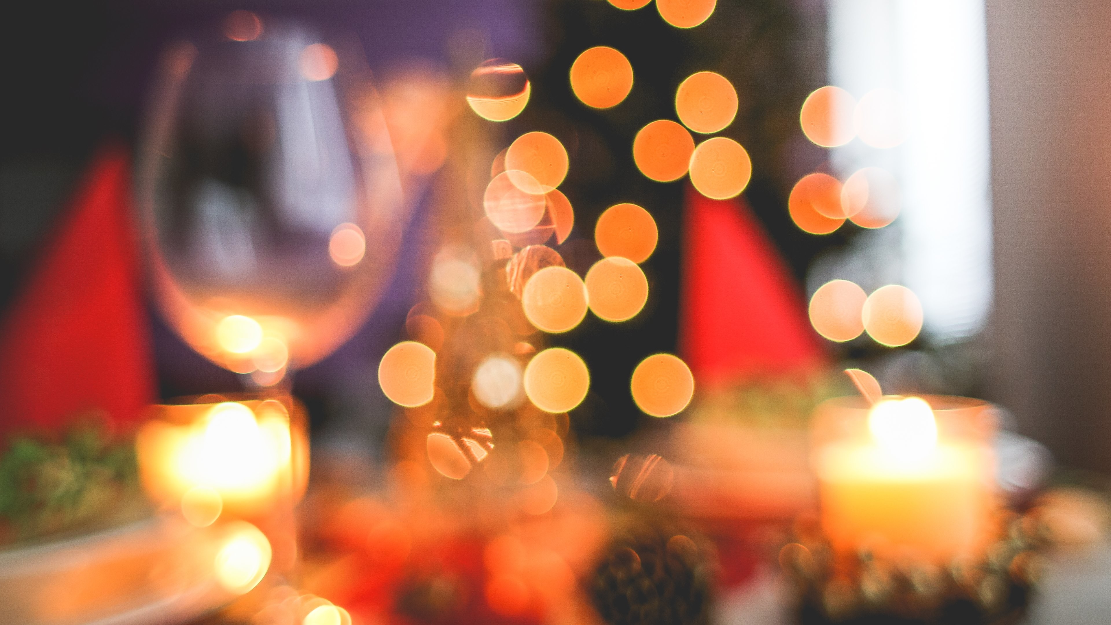 Blurred close-up of a festive Christmas table setting with a wine glass, candles, and bokeh lights from a Christmas tree in the background, creating a warm and inviting holiday atmosphere