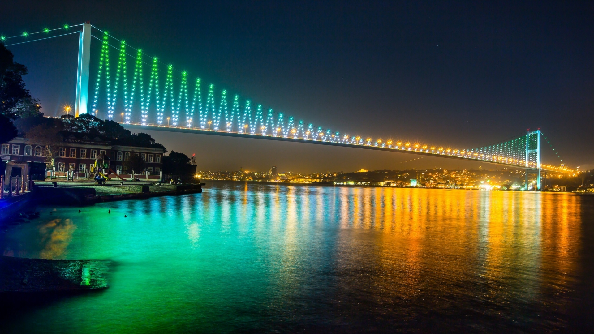 A breathtaking night view of the Yavuz Sultan Selim Bridge in Istanbul, illuminated with vibrant green and gold lights, reflecting beautifully on the Bosphorus