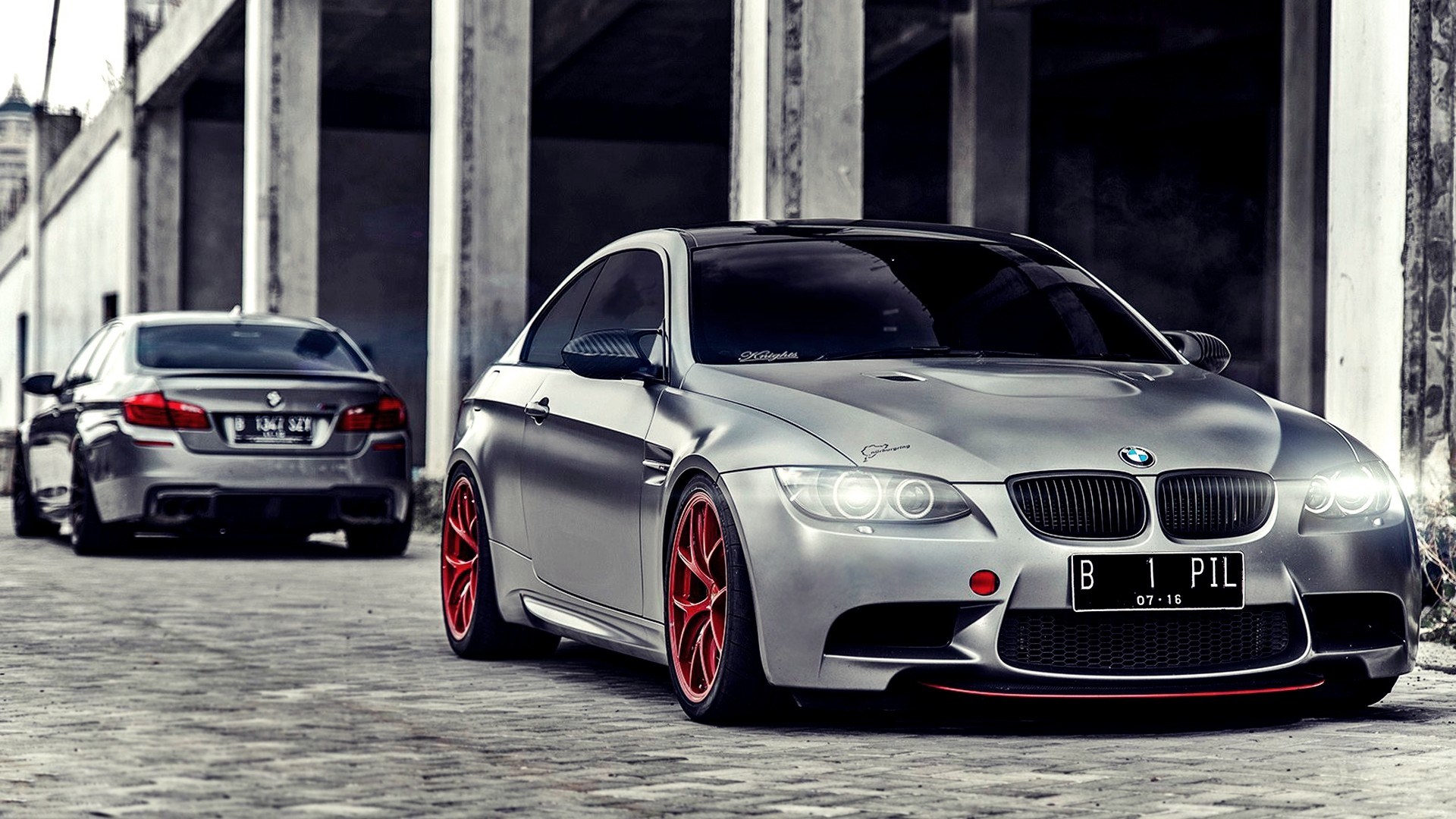 A metallic grey BMW M3 with red accents parked in a concrete structure, with a slightly blurred BMW 5 Series in the background