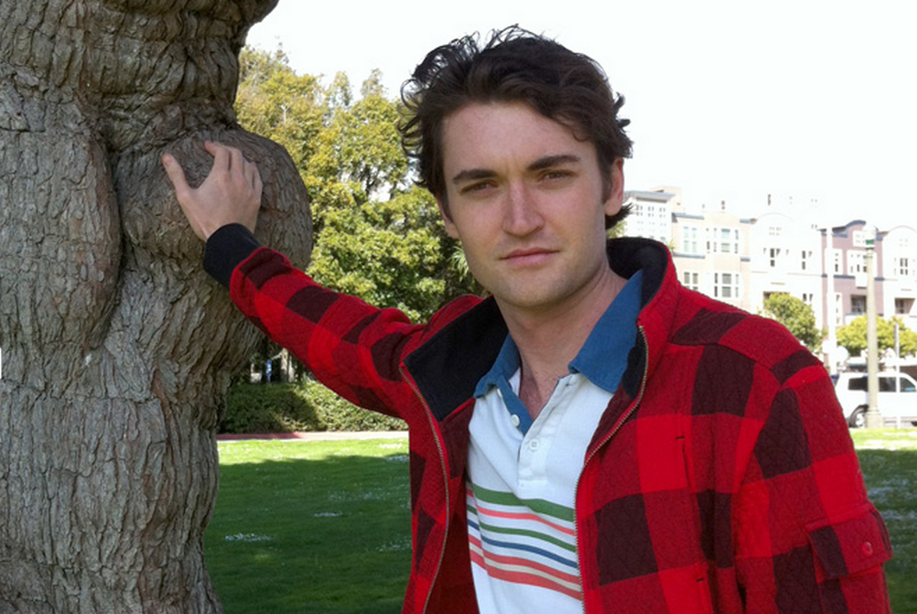 A person wearing a red and black plaid jacket, touching a tree trunk