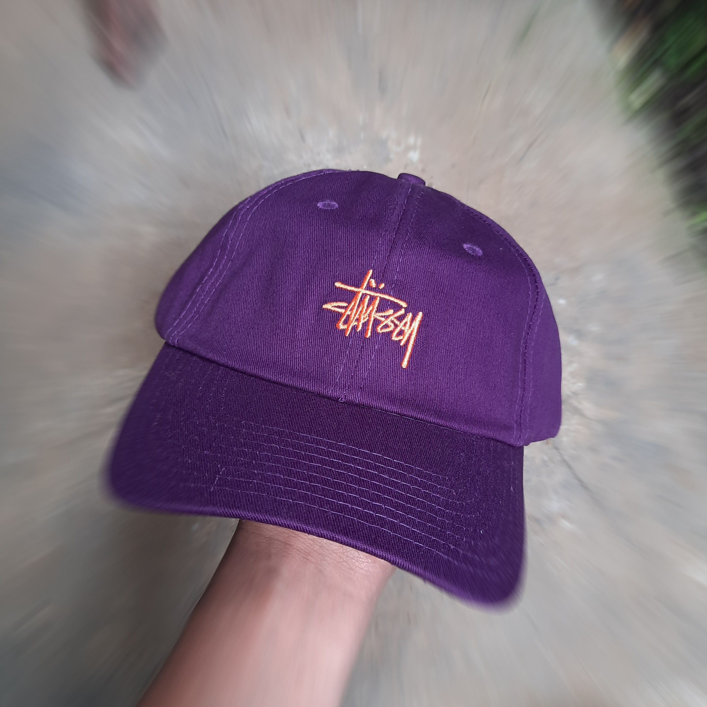 Close-up of a purple Stussy baseball cap held in hand, showcasing the embroidered Stussy logo in orange. The background has motion blur