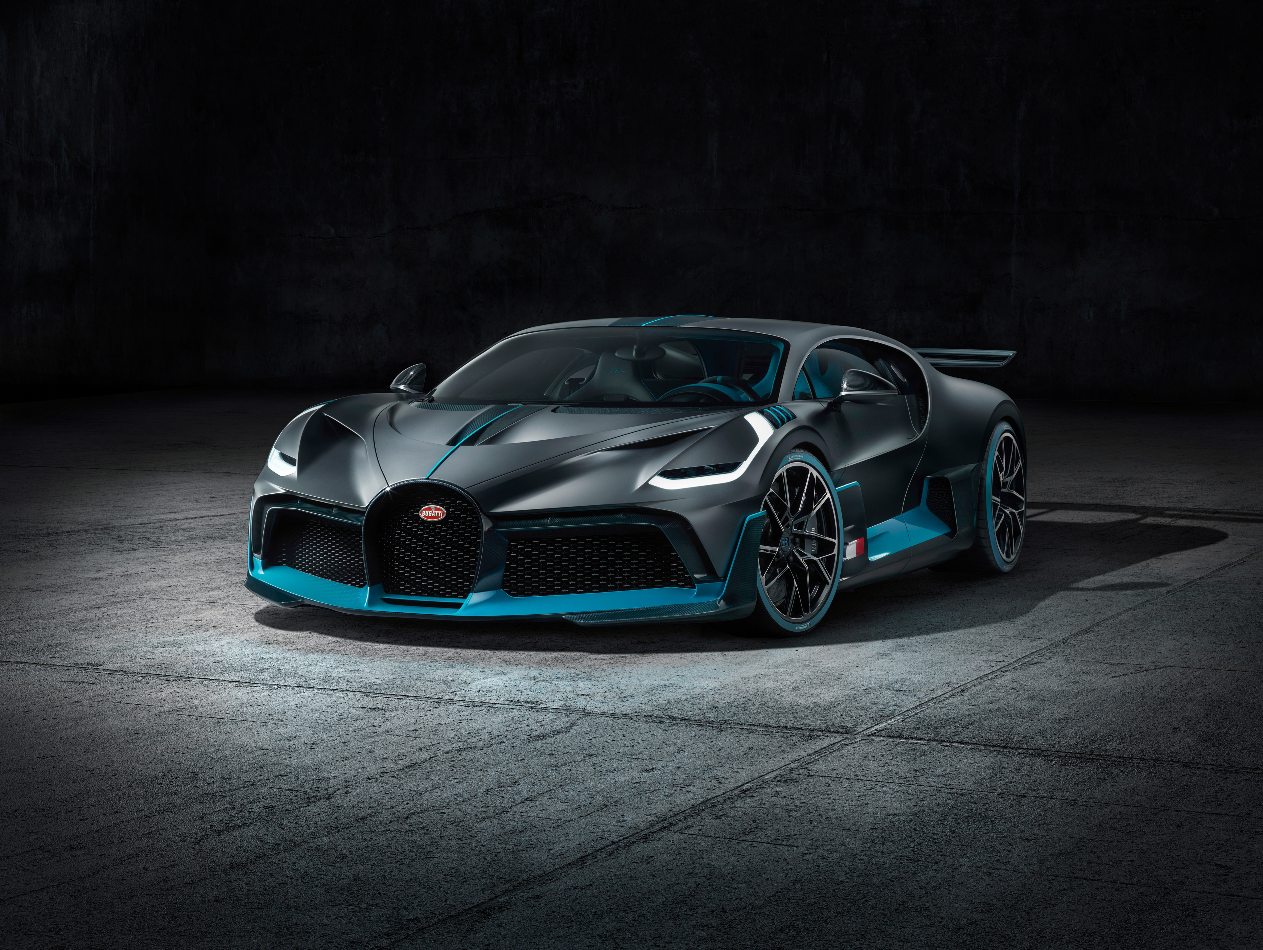 A sleek, dark gray Bugatti hypercar with vibrant teal accents, parked in a dimly lit industrial setting