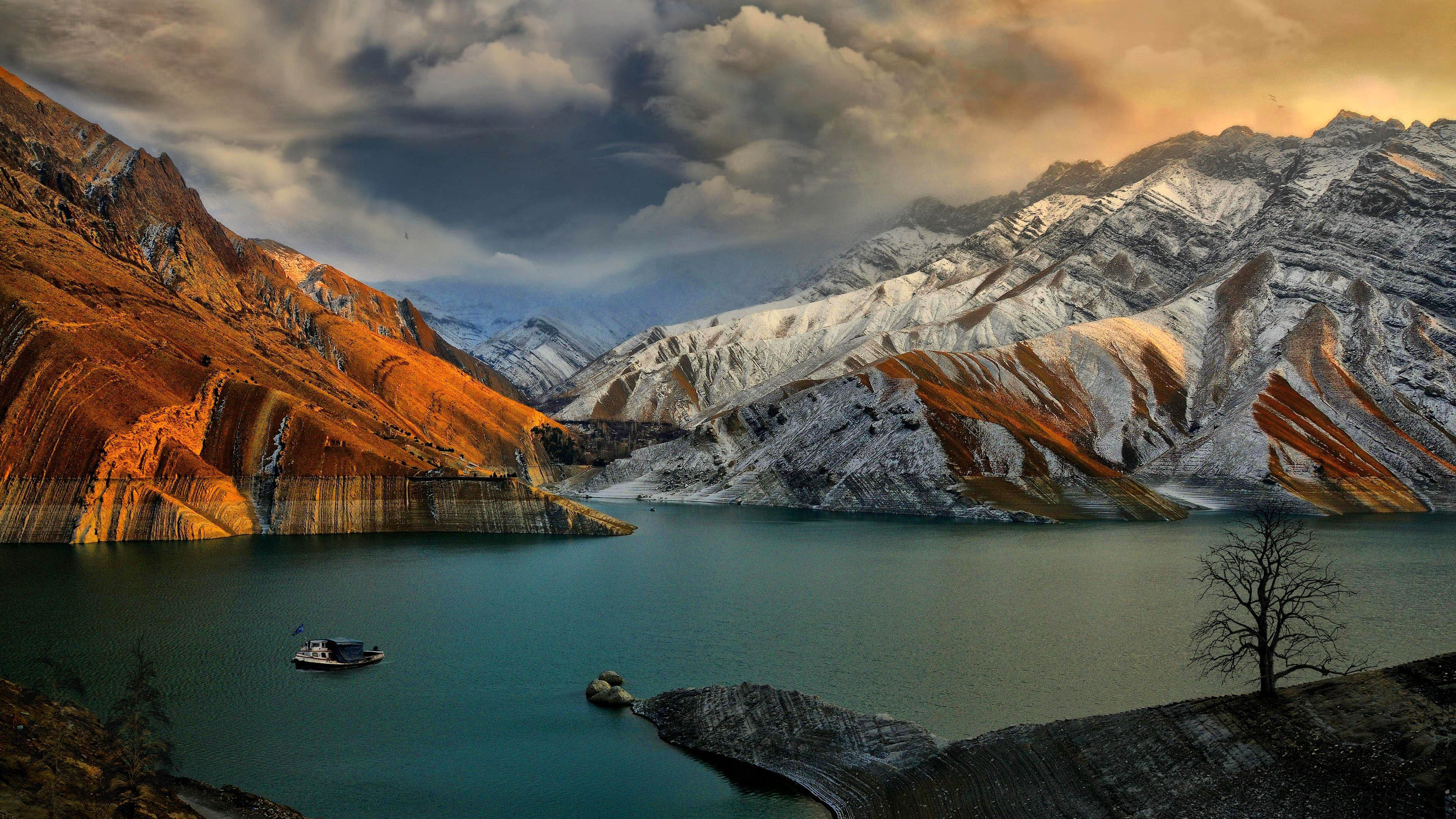 Stunning 4K background image of a serene lake nestled amidst snow-capped mountains at sunset, with a lone boat and bare tree adding to the tranquil ambiance