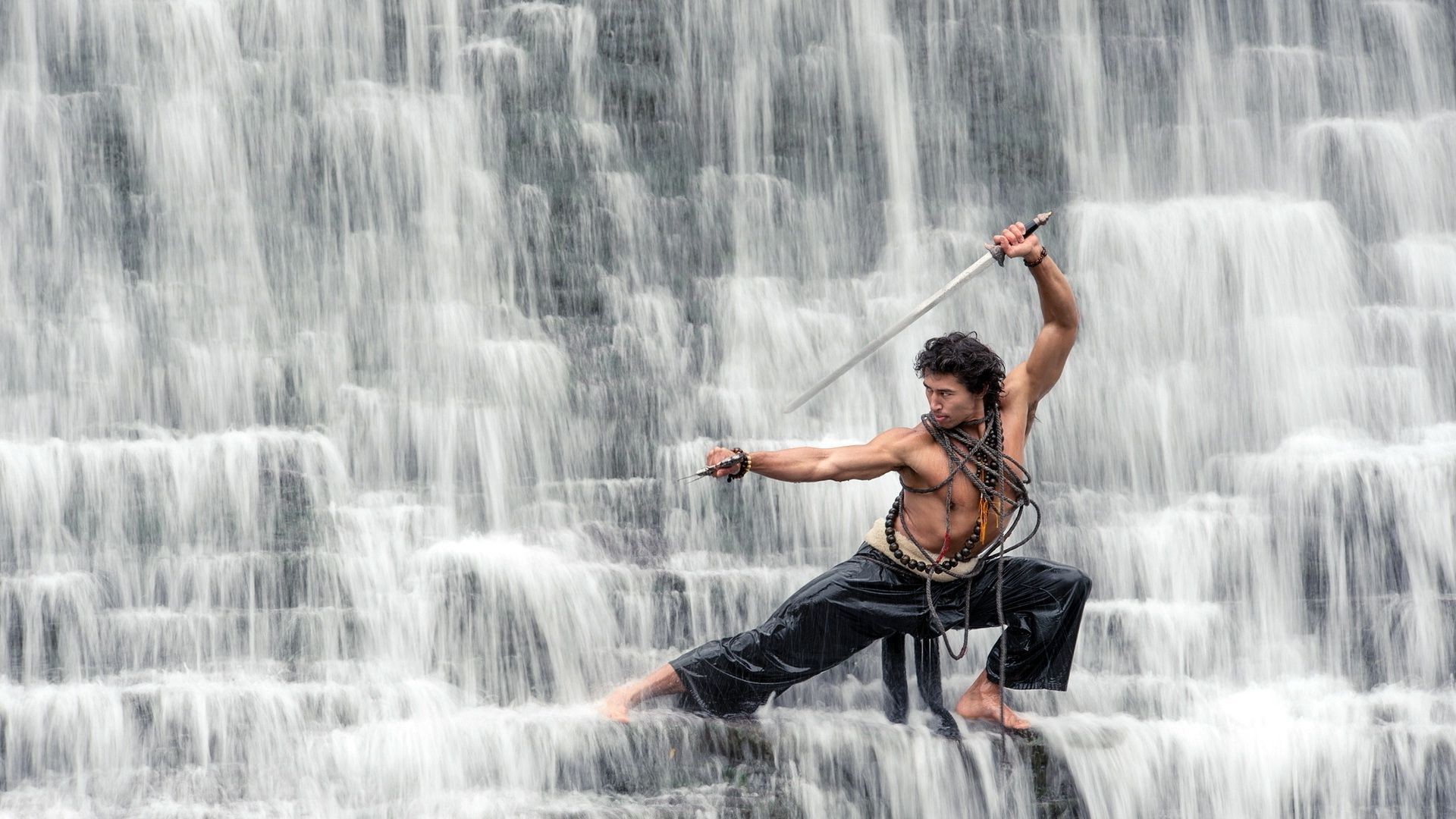 A martial artist strikes a dramatic pose with a sword in front of a cascading waterfall