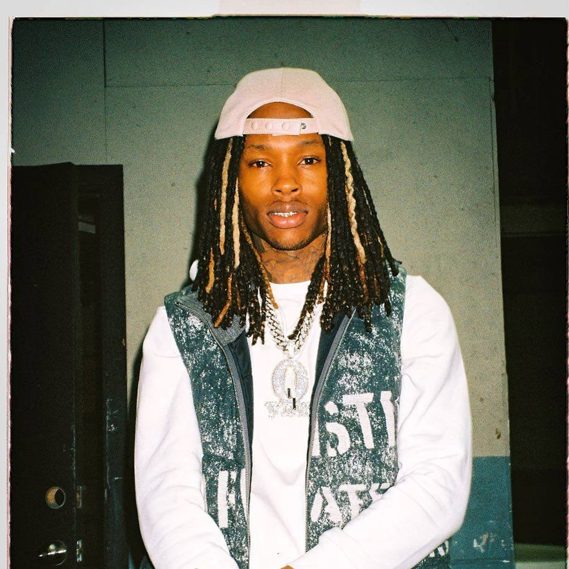 A portrait of a young man with dreadlocks, wearing a baseball cap, vest, white shirt, and multiple necklaces with a large pendant