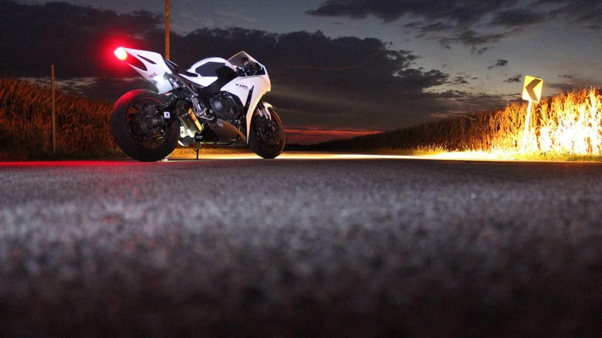 A white Honda CBR sportbike parked on a rural road at dusk, with its taillight glowing red and a curve sign illuminated in the background