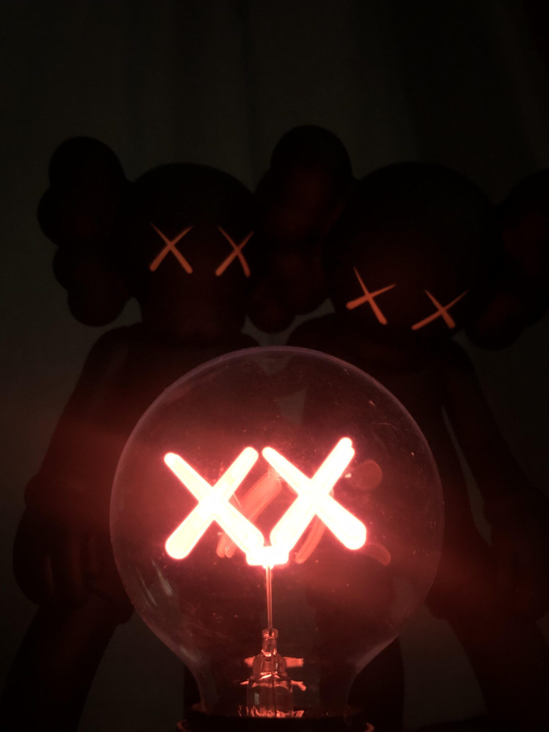 A glowing lightbulb featuring the signature XX of KAWS, positioned in front of two blurred KAWS Companion figures. This evokes a dark, artistic ambiance ideal for a desktop wallpaper
