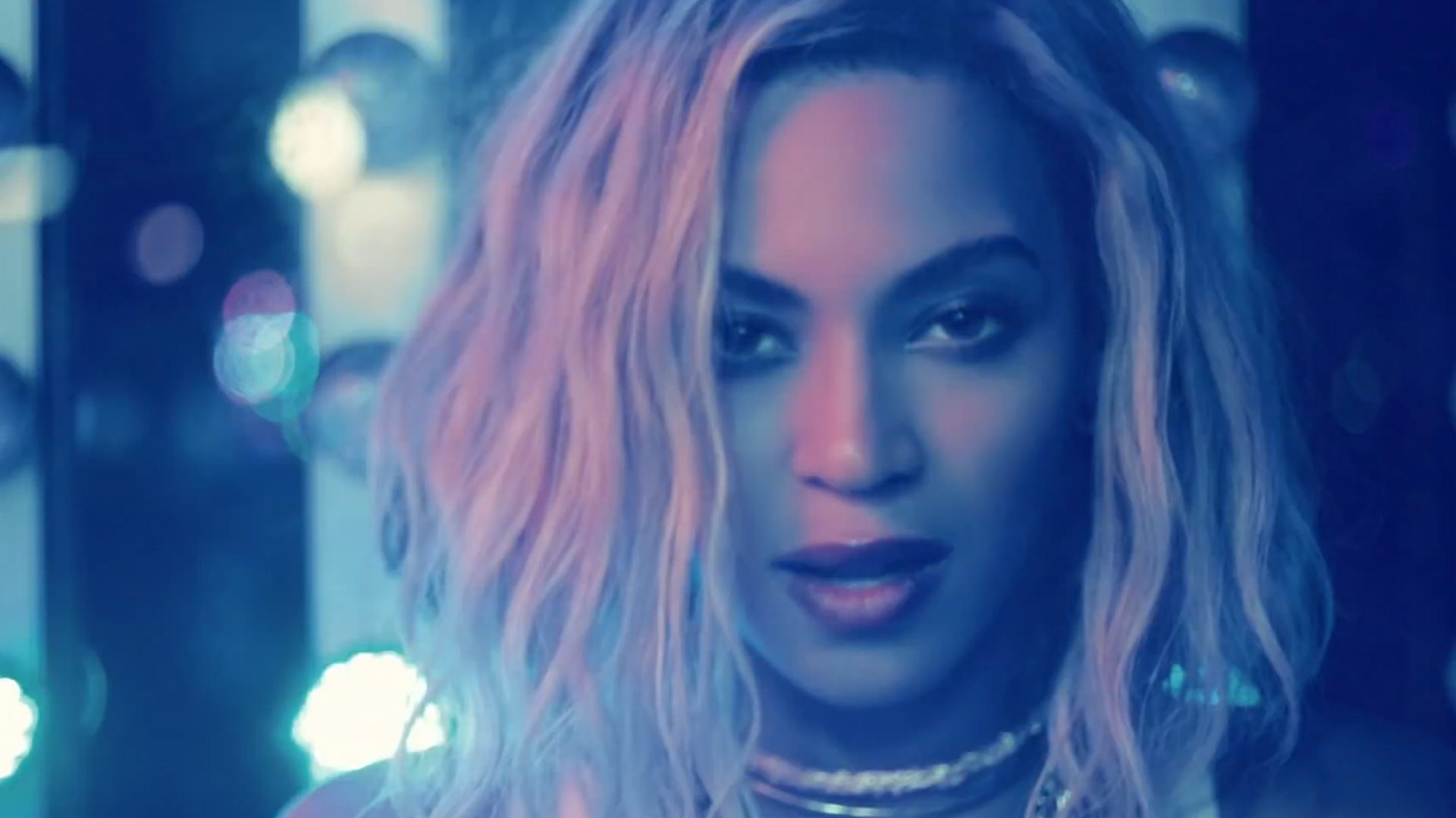 Close-up of Beyoncé with blonde hair, illuminated by blue and pink lighting, against a blurred background of lights