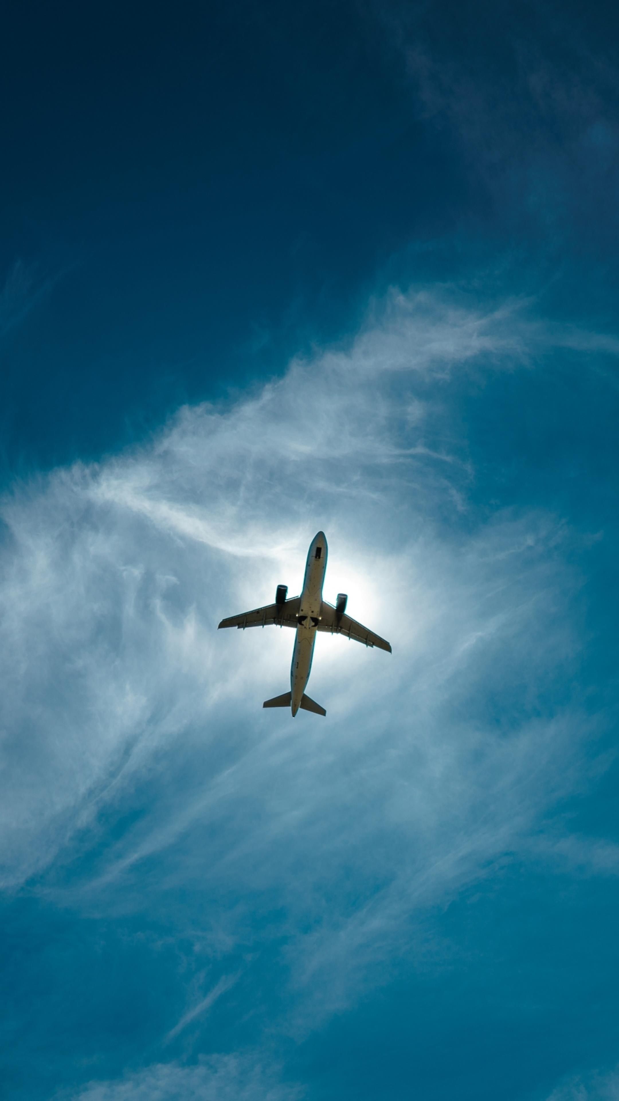 A passenger airplane soars high in the vibrant blue sky, slightly obscured by wispy white clouds. Its sleek silhouette is backlit by the sun, making it appear almost as a shadow against the backdrop of the sky
