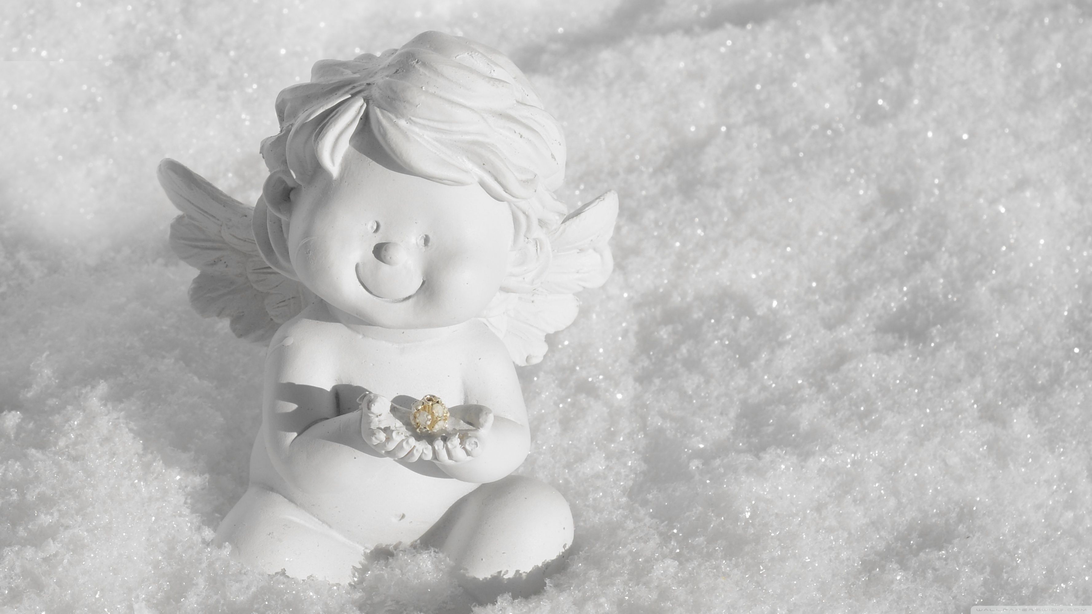 A small white angel figurine sits in the snow, holding a delicate gold and white snowflake ornament. Its serene expression and the pristine snow create a peaceful winter scene