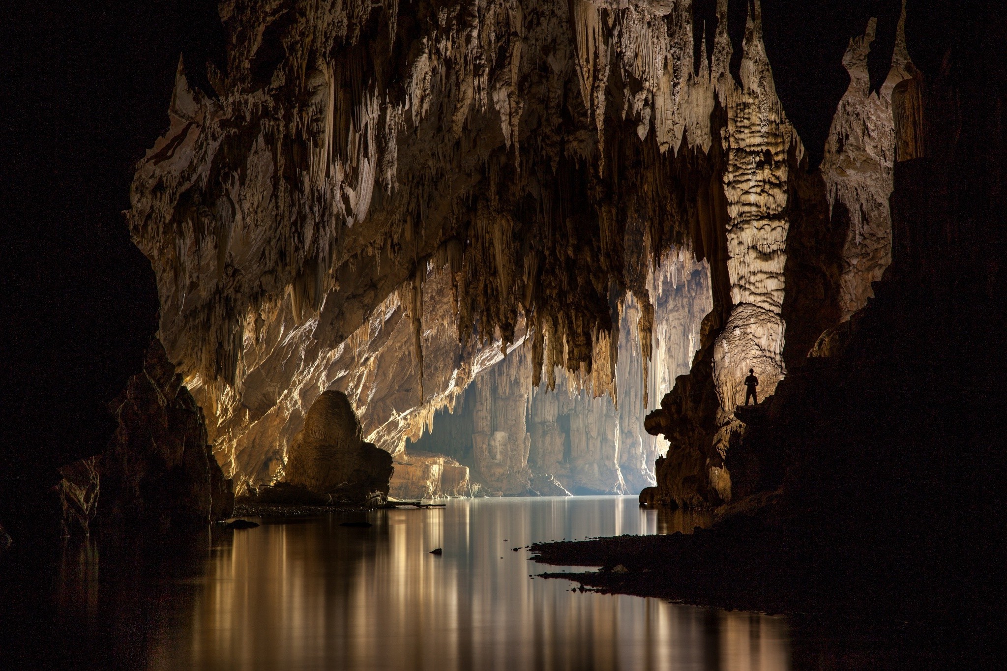 A breathtaking view of an underground cave with stalactites hanging from the ceiling and a calm body of water reflecting the cave's formations. A lone explorer stands in silhouette, emphasizing the vastness and mystery of the cavern