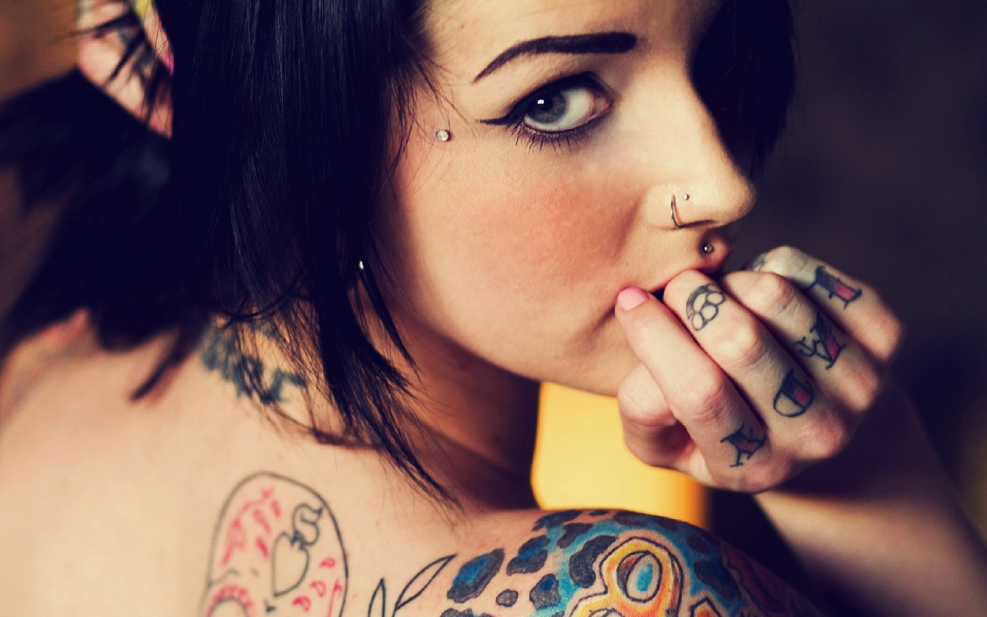 Close-up of a woman with multiple tattoos, including a sugar skull on her back and lettering on her fingers, and several piercings