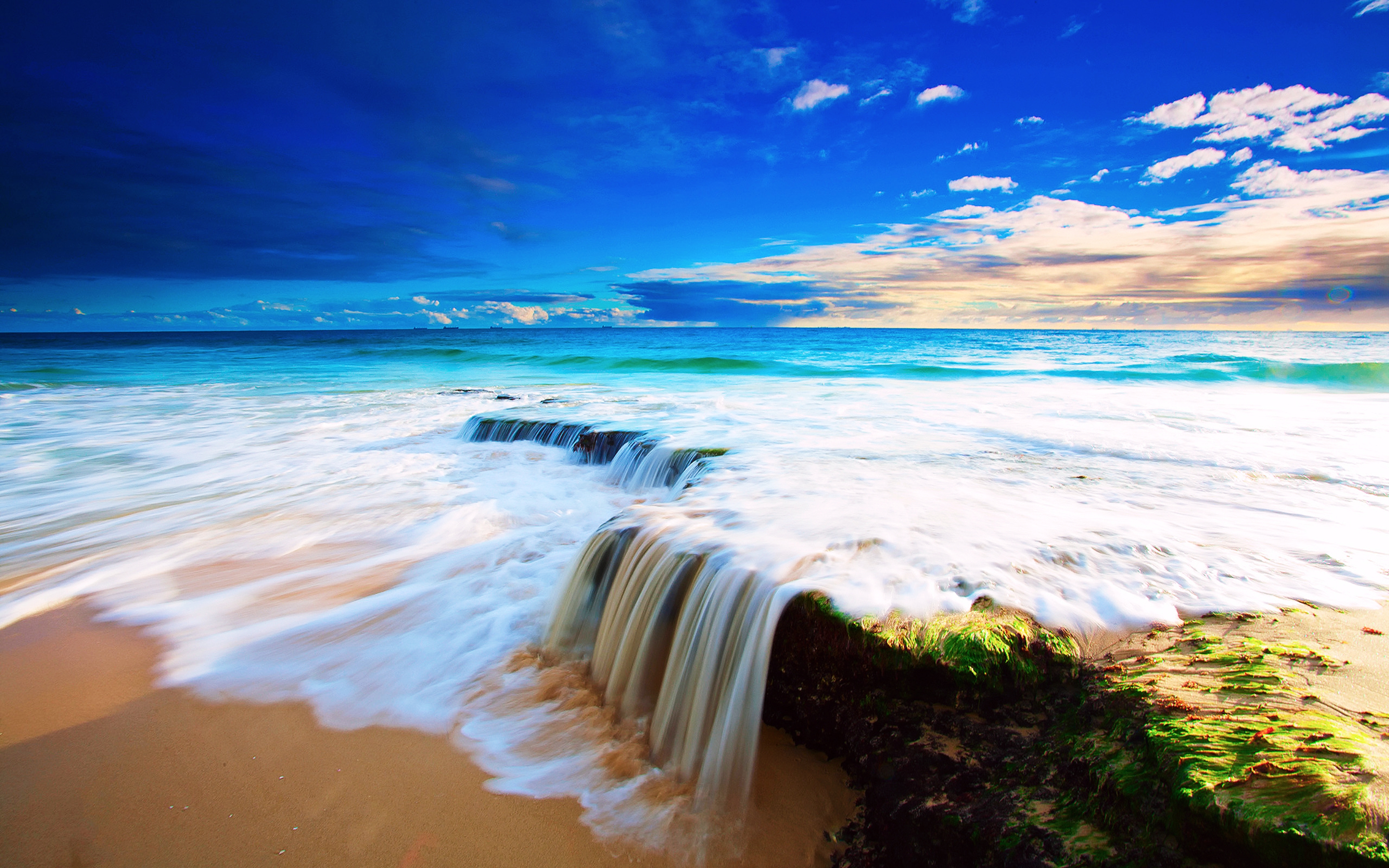 A cascading waterfall flows onto a sandy beach, with the vibrant turquoise ocean and a colorful sky in the background