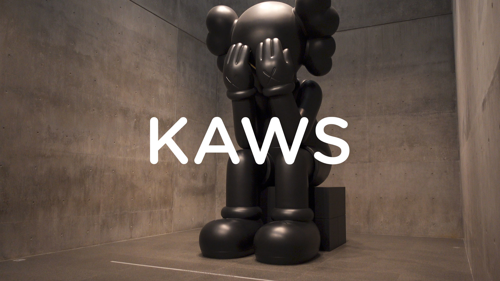 A large black KAWS Companion sculpture sits on a plinth in a concrete room, its hands covering its face. This iconic piece showcases the artist's signature style