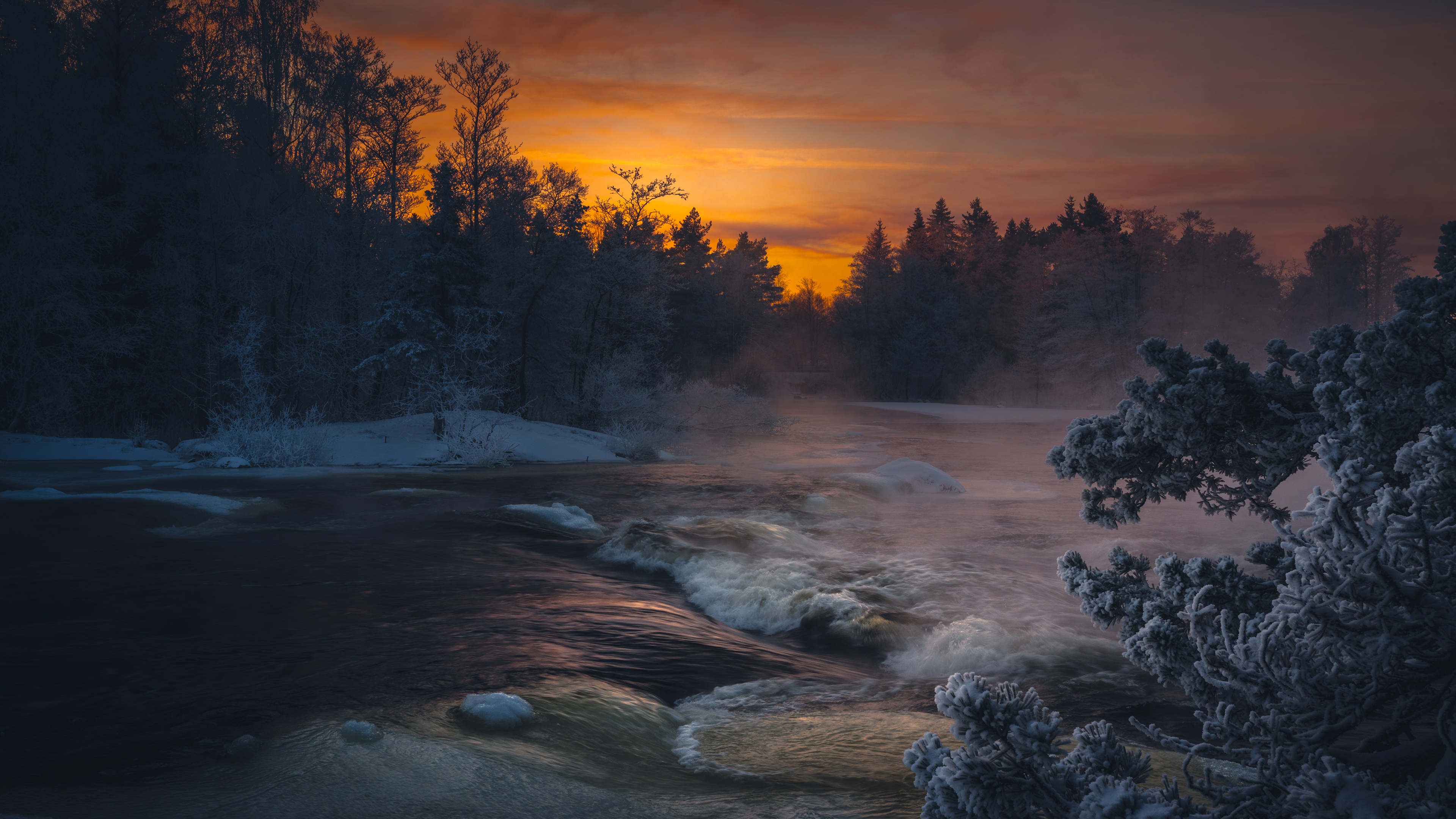 A breathtaking 4K wallpaper of a winter river at sunset. The icy river flows through a snowy forest, reflecting the warm hues of the setting sun. Frost-covered trees frame the scene, creating a serene and captivating winter landscape