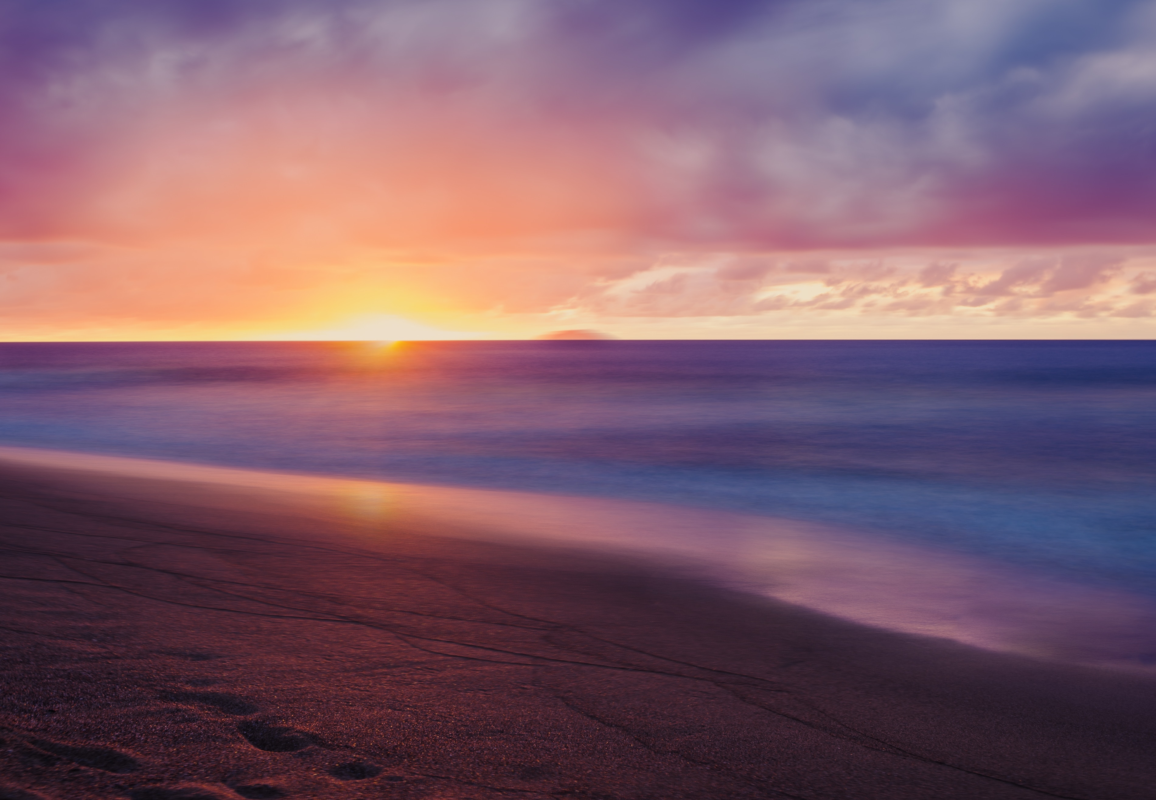A breathtaking 4K wallpaper capturing the serene beauty of a beach at sunset. The vibrant hues of orange, pink, and purple paint the sky as the sun dips below the horizon, casting a warm glow on the smooth, sandy shore and the gentle waves lapping at the edge