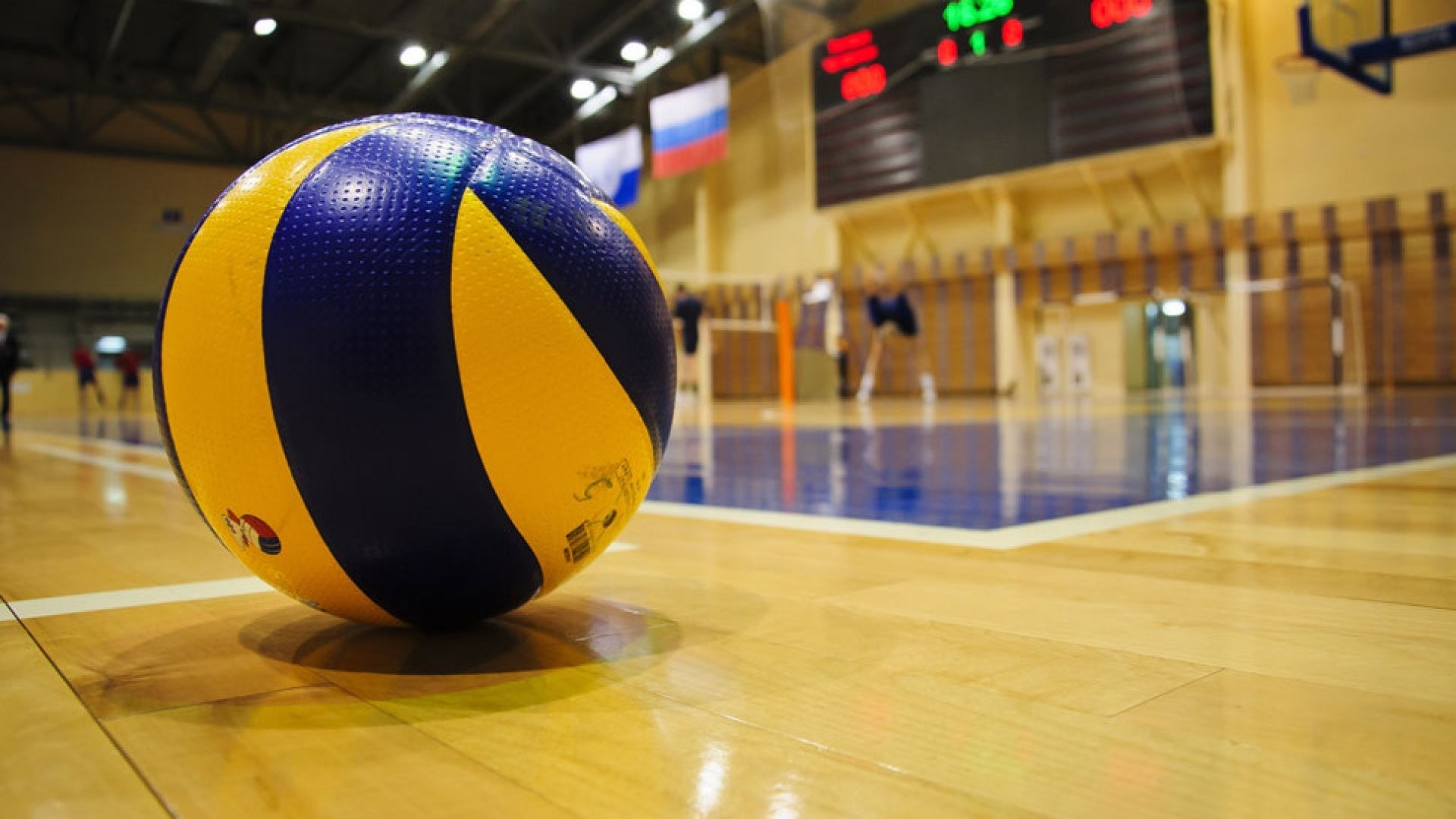 A close-up of a volleyball on a polished wooden court, with blurred players in the background, ready for a serve