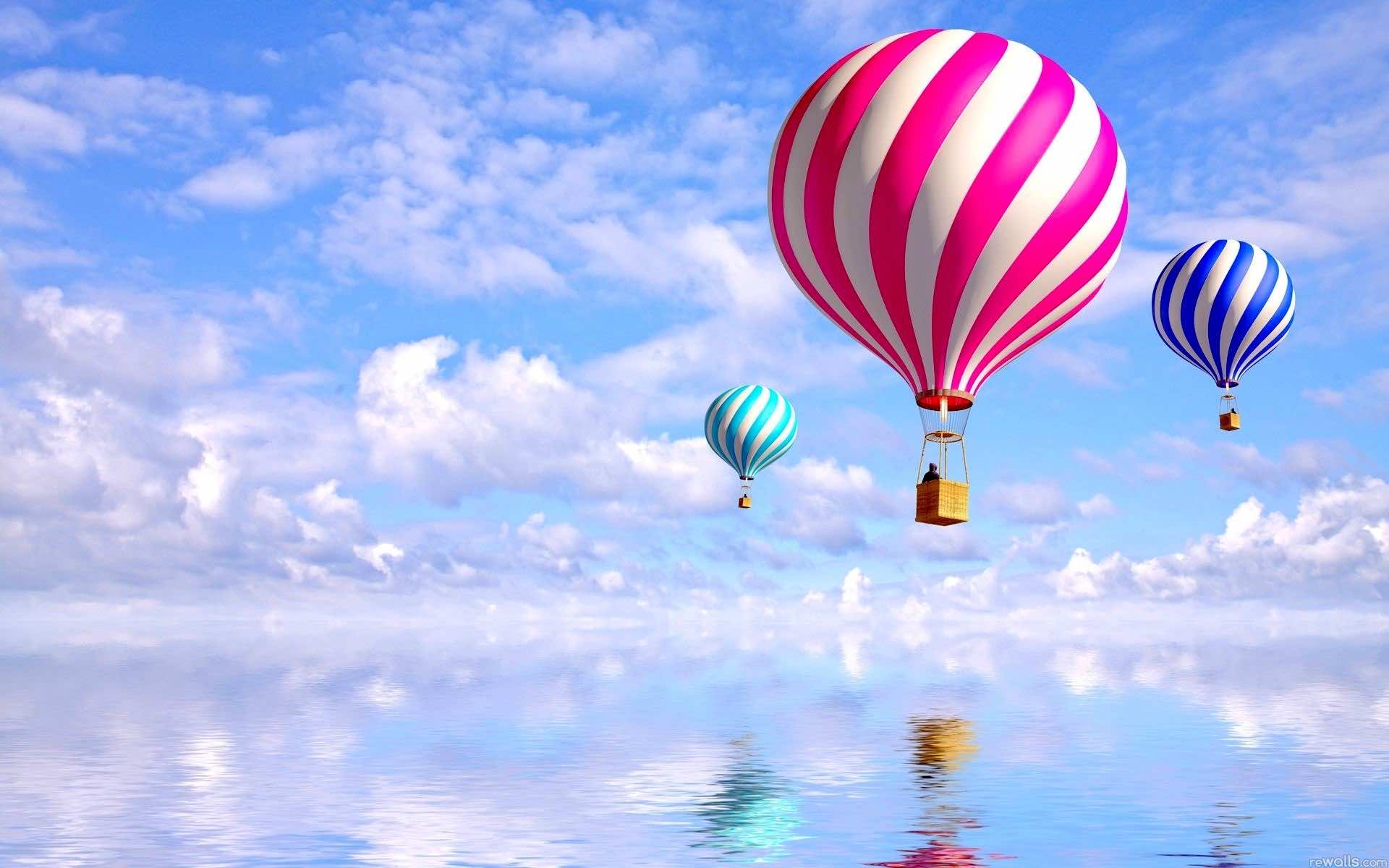 Three hot air balloons soaring over a calm body of water, reflecting the sky and clouds