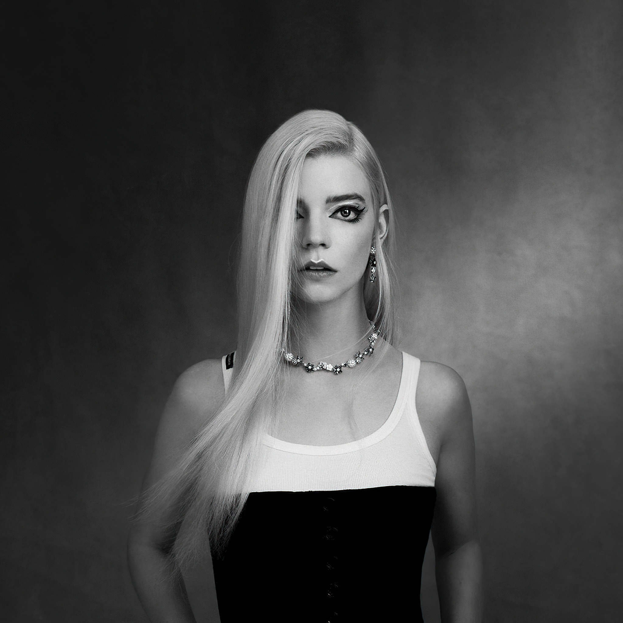 Black and white portrait of Anya Taylor-Joy with long blonde hair, wearing a white tank top and black corset, against a dark background