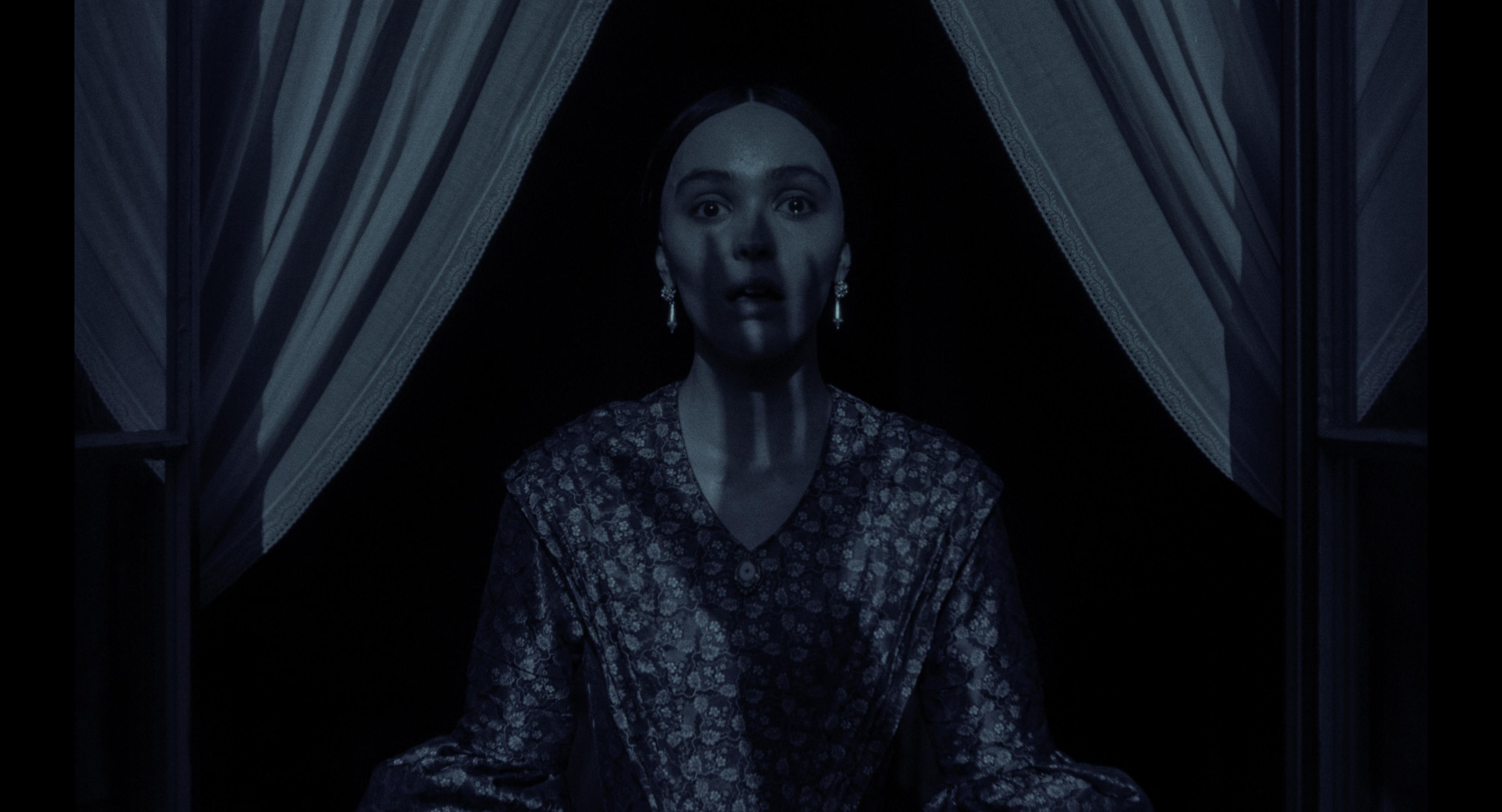 A woman with a fearful expression, possibly from the movie 'Nosferatu 2024', stands in a dimly lit room, partially obscured by curtains. A shadowy handprint is cast across her face, adding to the suspenseful atmosphere