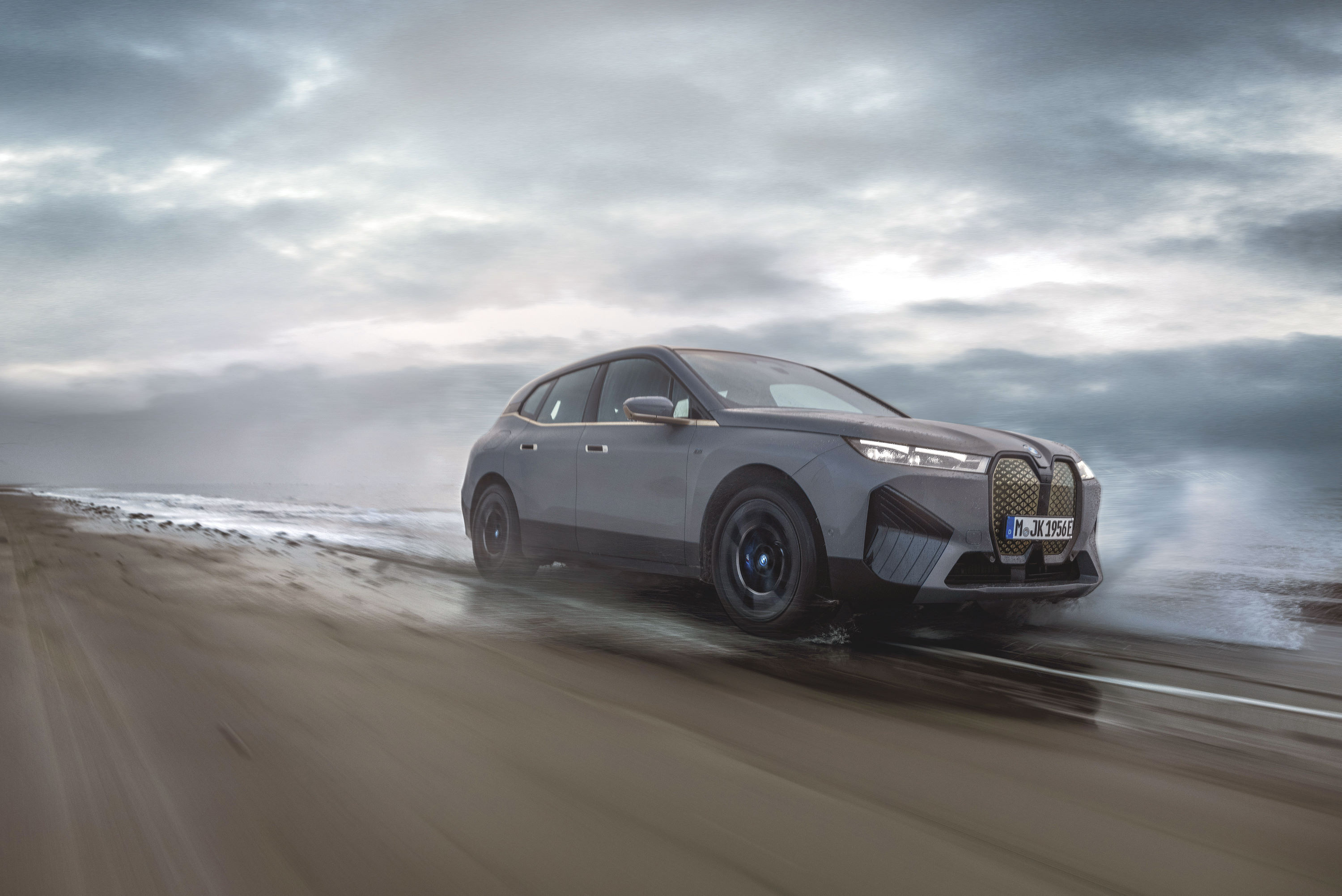 A BMW iX electric SUV driving on a wet, sandy beach against a dramatic cloudy sky