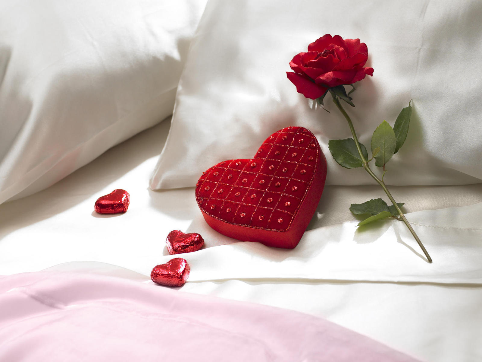 Romantic still life of a red rose, heart-shaped box, and chocolate hearts on a bed with white and pink satin sheets