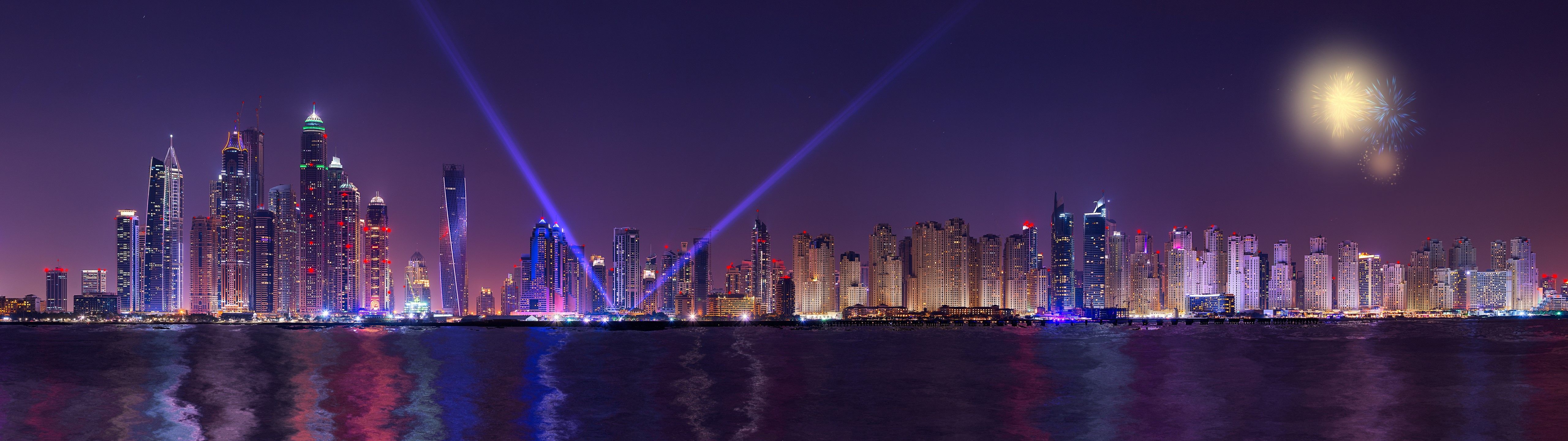 Ultrawide panoramic view of Dubai Marina skyscrapers illuminated at night with searchlights and fireworks, reflecting in the water