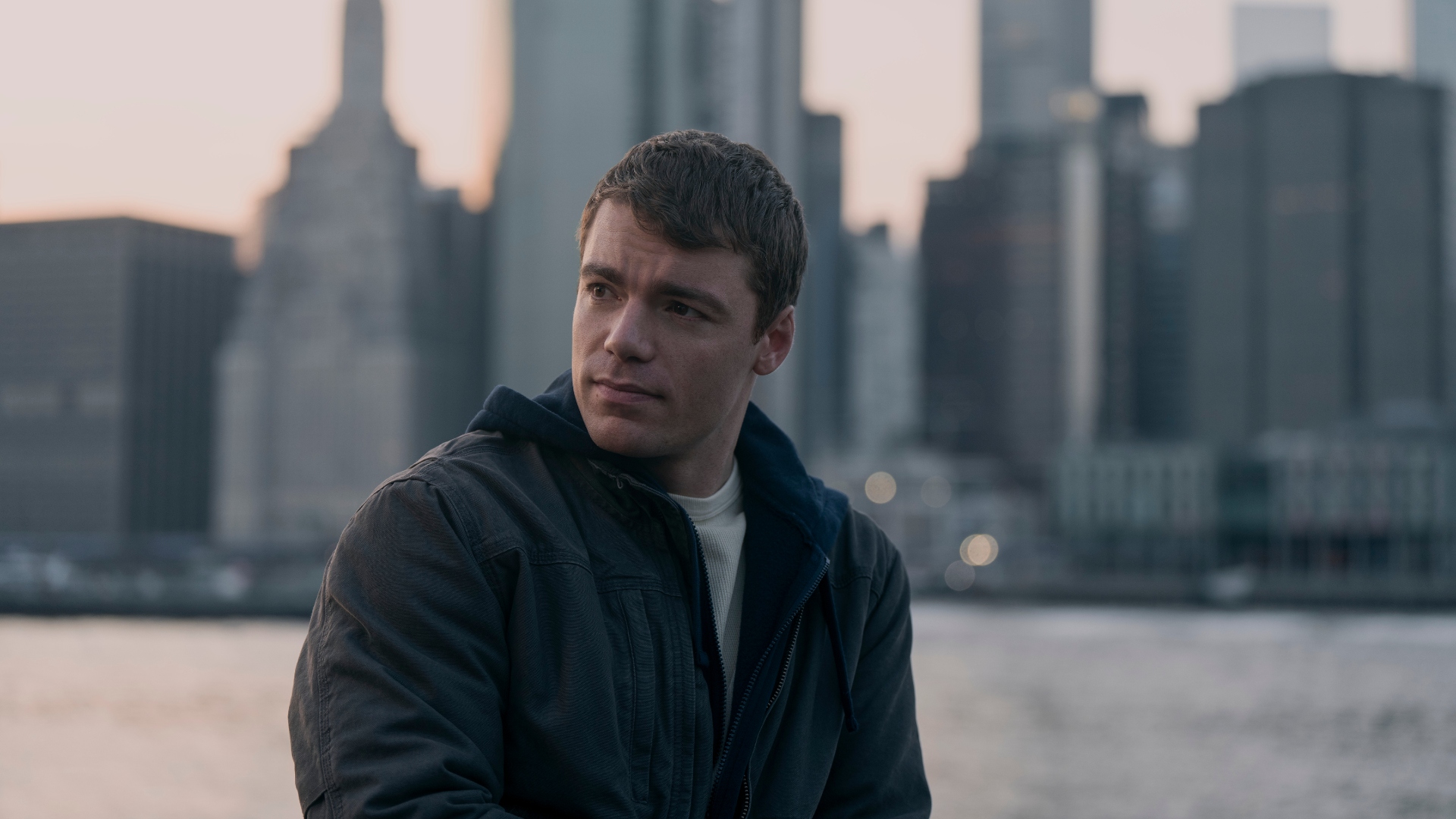 A young man in a dark jacket stands against a blurred cityscape backdrop at sunset