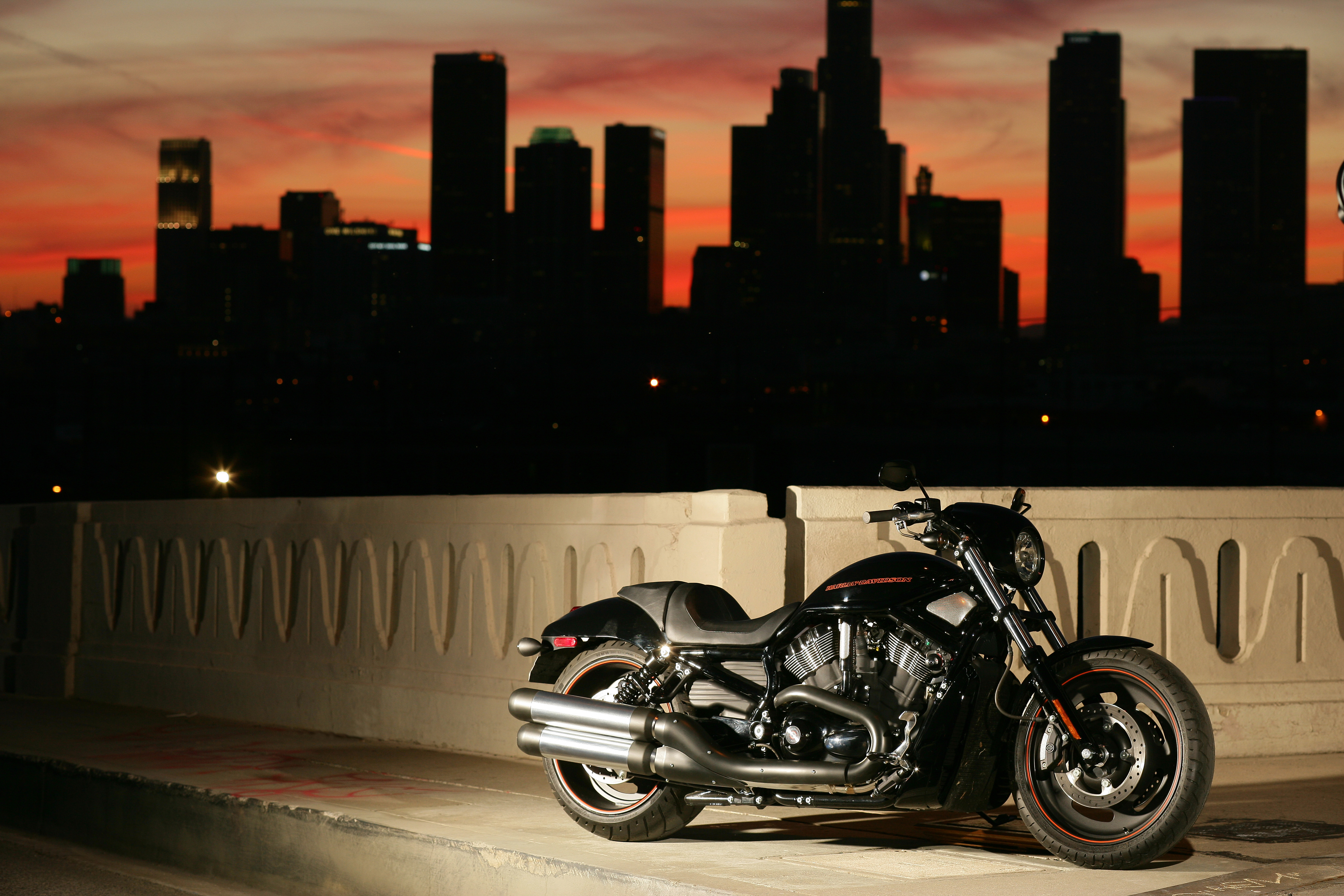 A black Harley-Davidson V-Rod Night Rod Special parked on a bridge at sunset with the Los Angeles skyline in the background