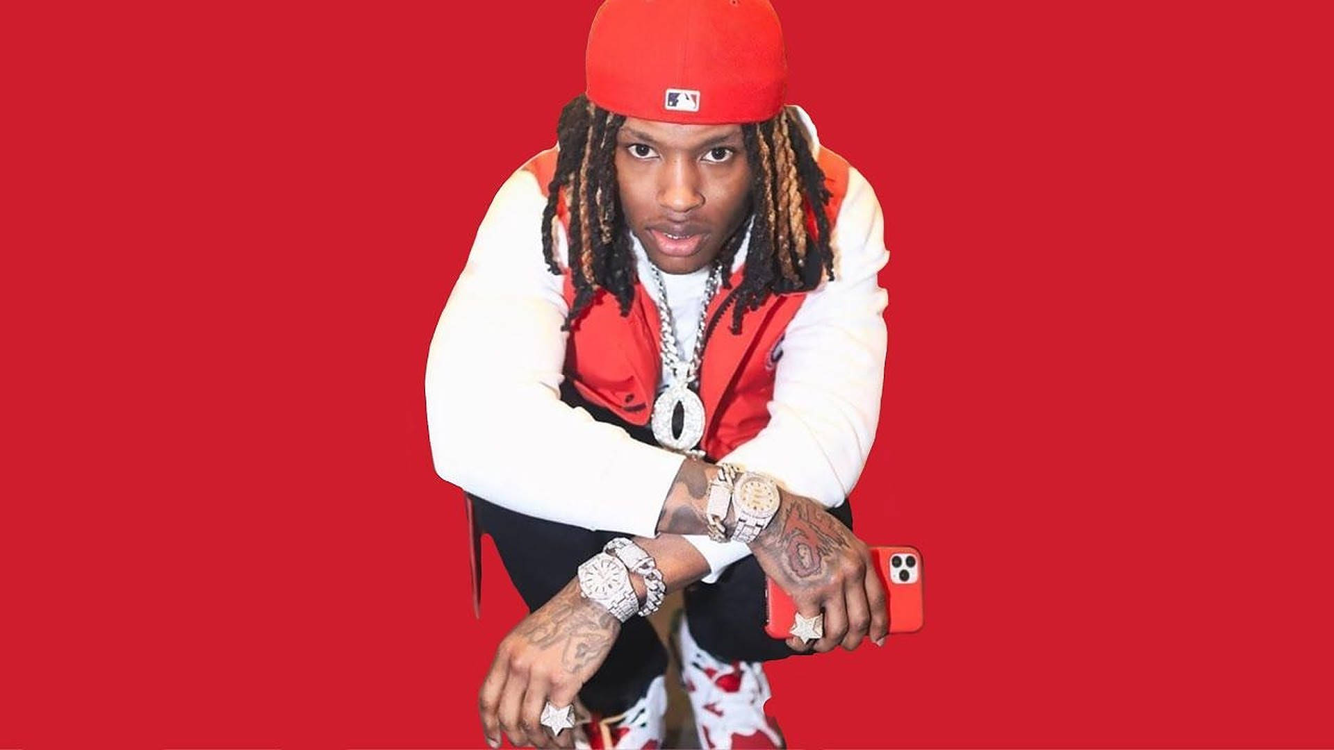 A rapper in a red baseball cap, red vest, white shirt, and iced-out jewelry poses against a red background