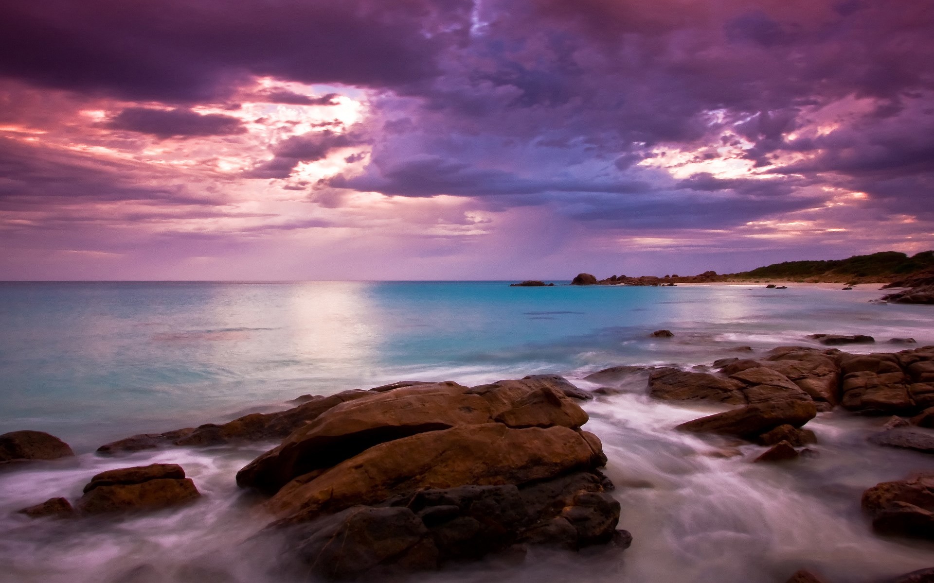 A breathtaking sunset over a rocky beach, with purple and pink hues illuminating the sky and turquoise waters gently lapping against the shore
