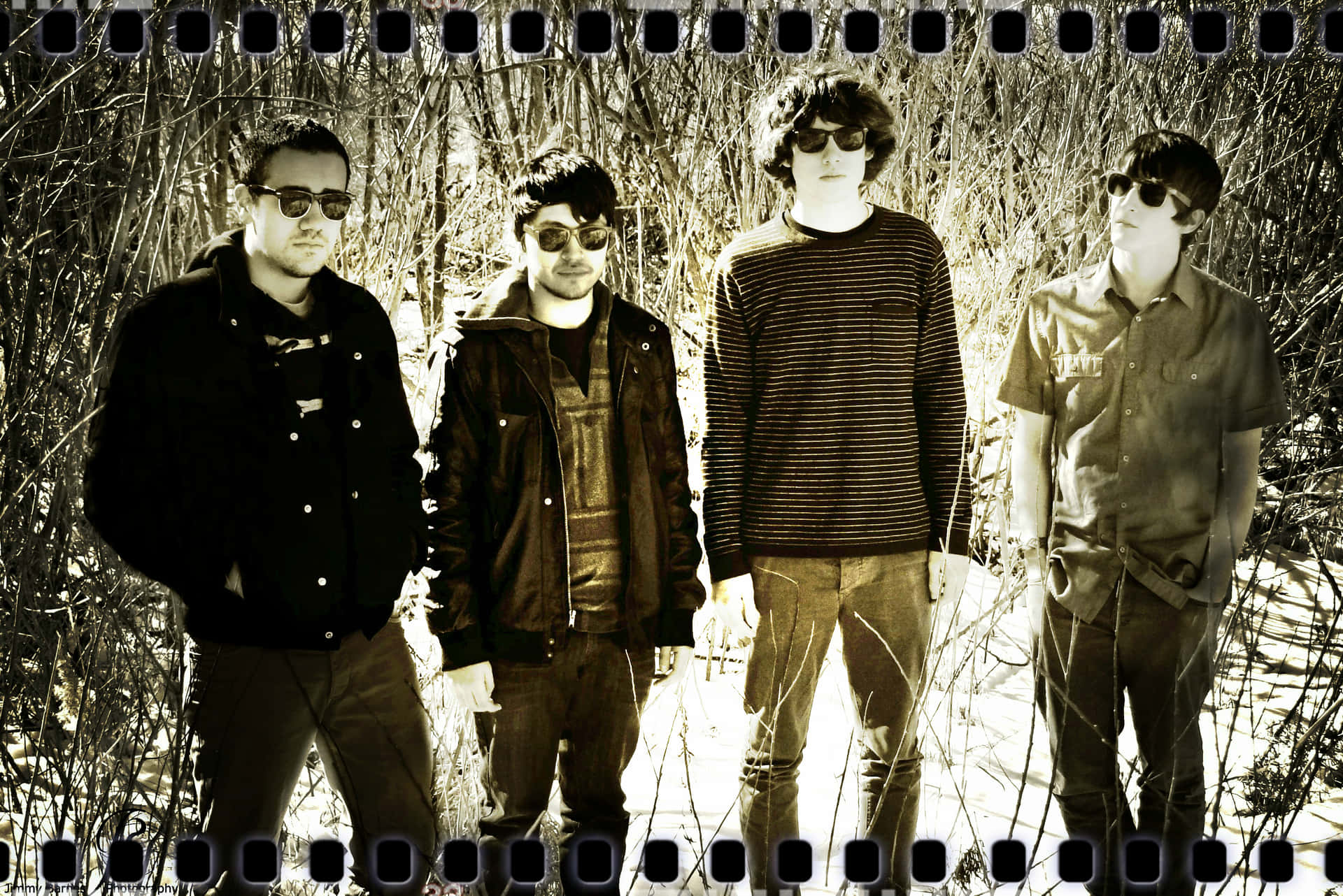 Four individuals pose for a photo in a snowy, wooded area. They are dressed in casual attire, including jackets, sweaters, and sunglasses