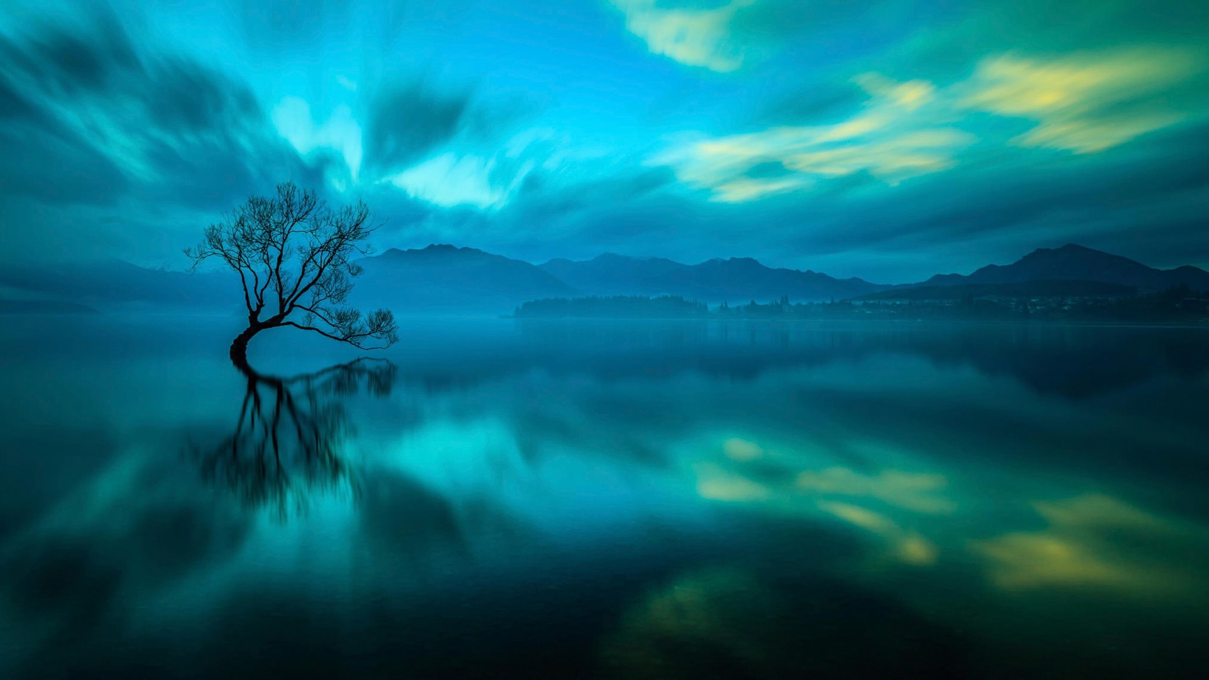 A lone tree stands in the serene waters of Lake Wanaka, New Zealand, its reflection mirrored in the tranquil surface. The background showcases a misty mountain range under a breathtaking, teal-colored sky. This stunning natural landscape captures the ethereal beauty of nature at its finest