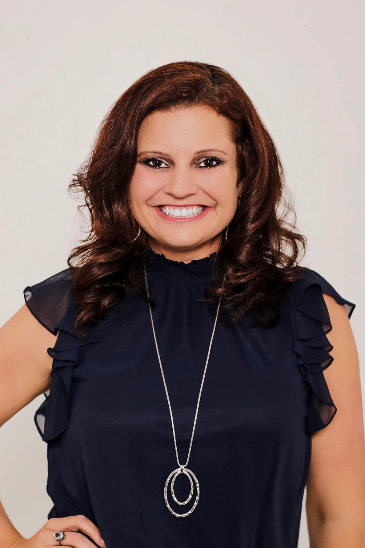 Headshot of a woman with shoulder-length auburn hair, smiling at the camera. She is wearing a navy blue blouse with ruffled sleeves and a silver pendant necklace
