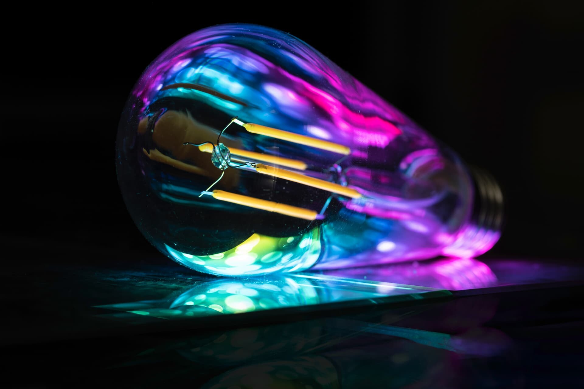 A multicolored LED light bulb lies on a reflective surface, casting vibrant hues of blue, green, and pink. The intricate filaments of the bulb are visible through the clear glass, creating a captivating interplay of light and shadow